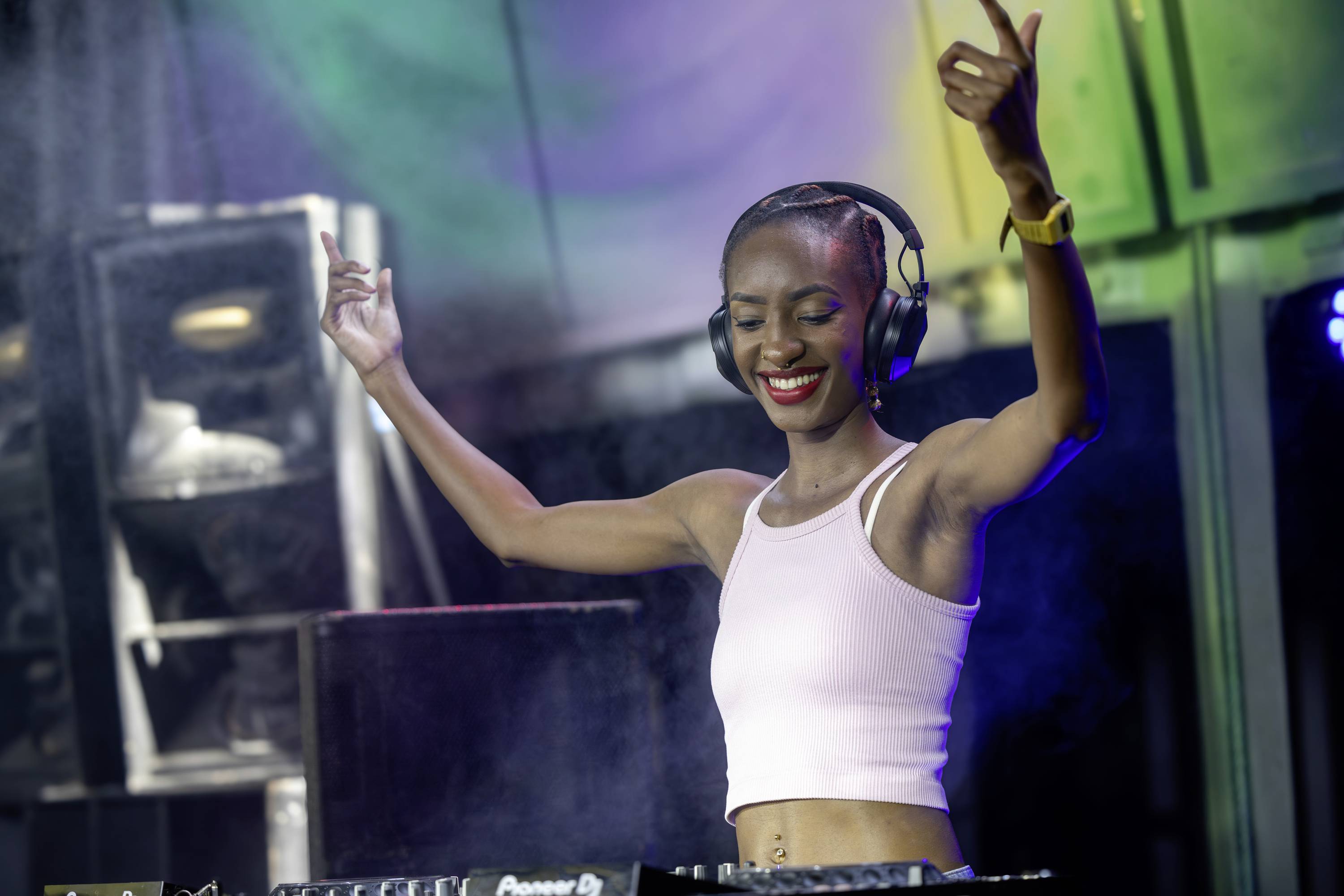 A DJ on a stage is wearing headphones and raising her arms in the air with a smile while playing music.