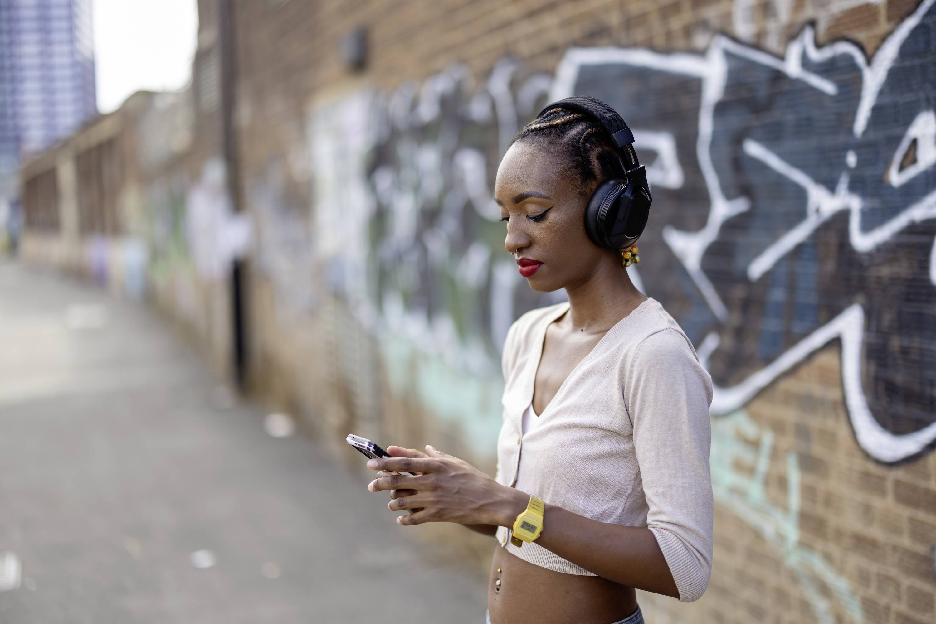 A woman wearing headphones is standing in front of a brick wall with graffiti, looking at her mobile phone. She is wearing a white blouse.