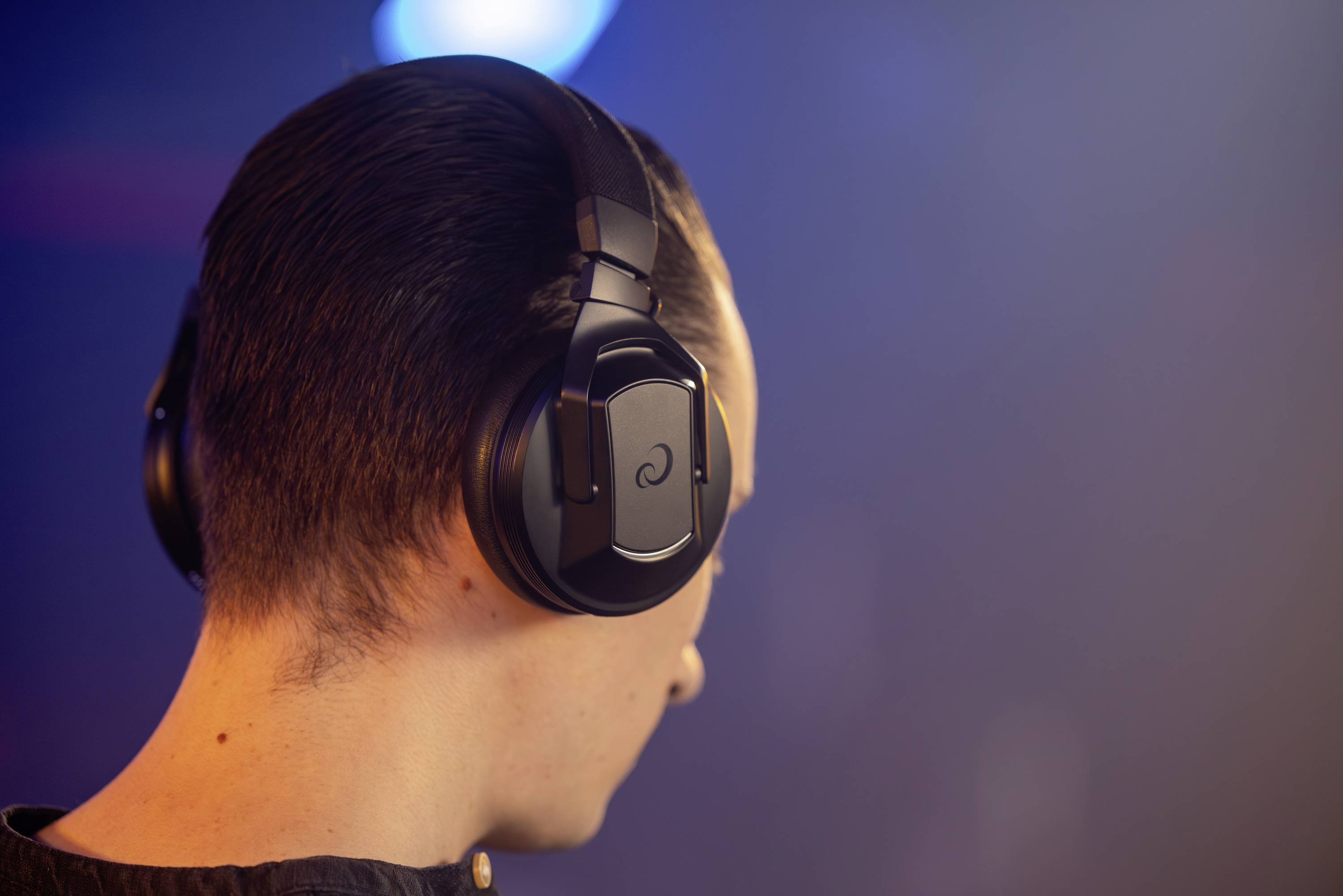 A person is wearing black, wireless headphones and listening to music. They are viewed from behind, with bright light in the background.