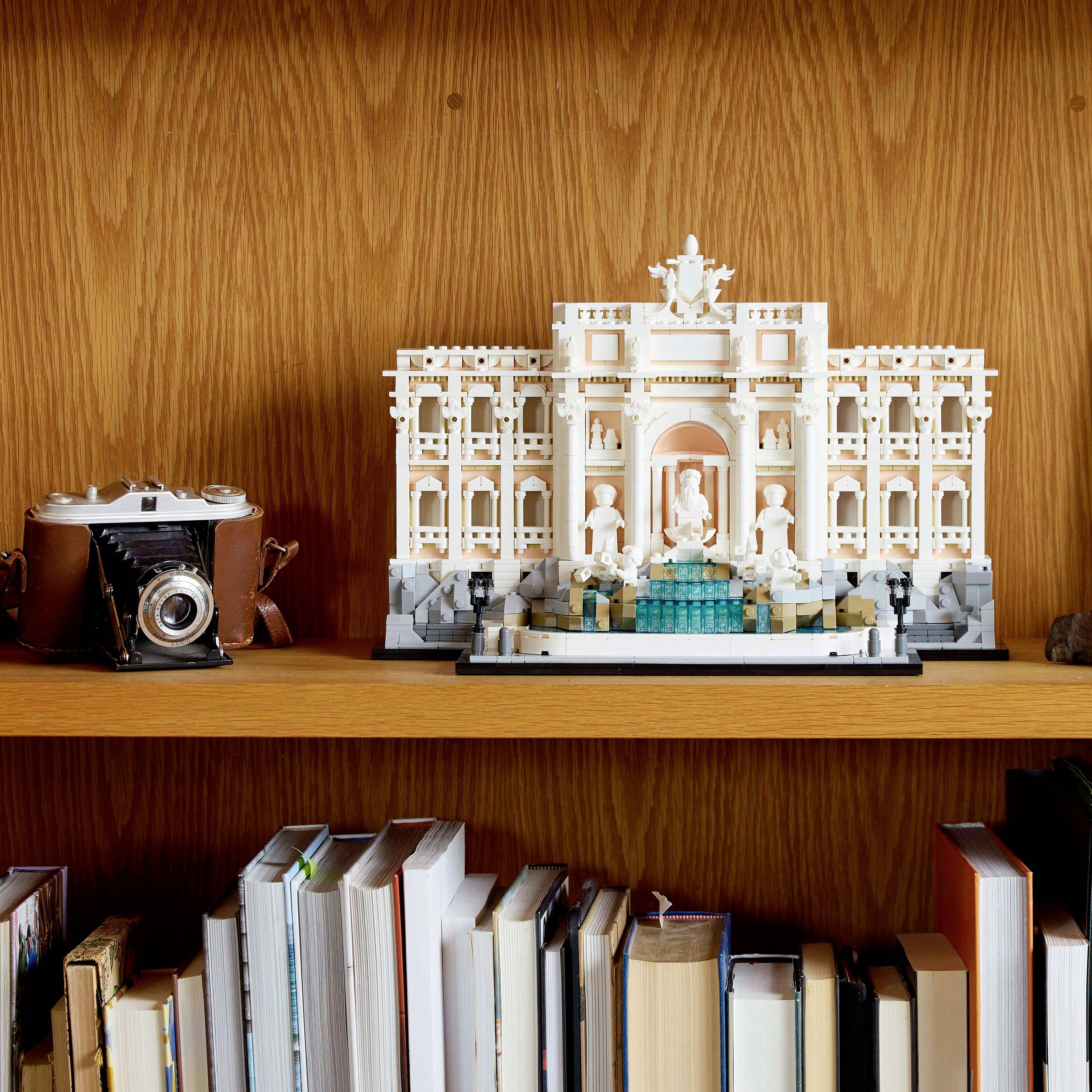 A Lego model of the Trevi Fountain sits on a wooden shelf. An old camera lies to the left, with books stacked underneath.