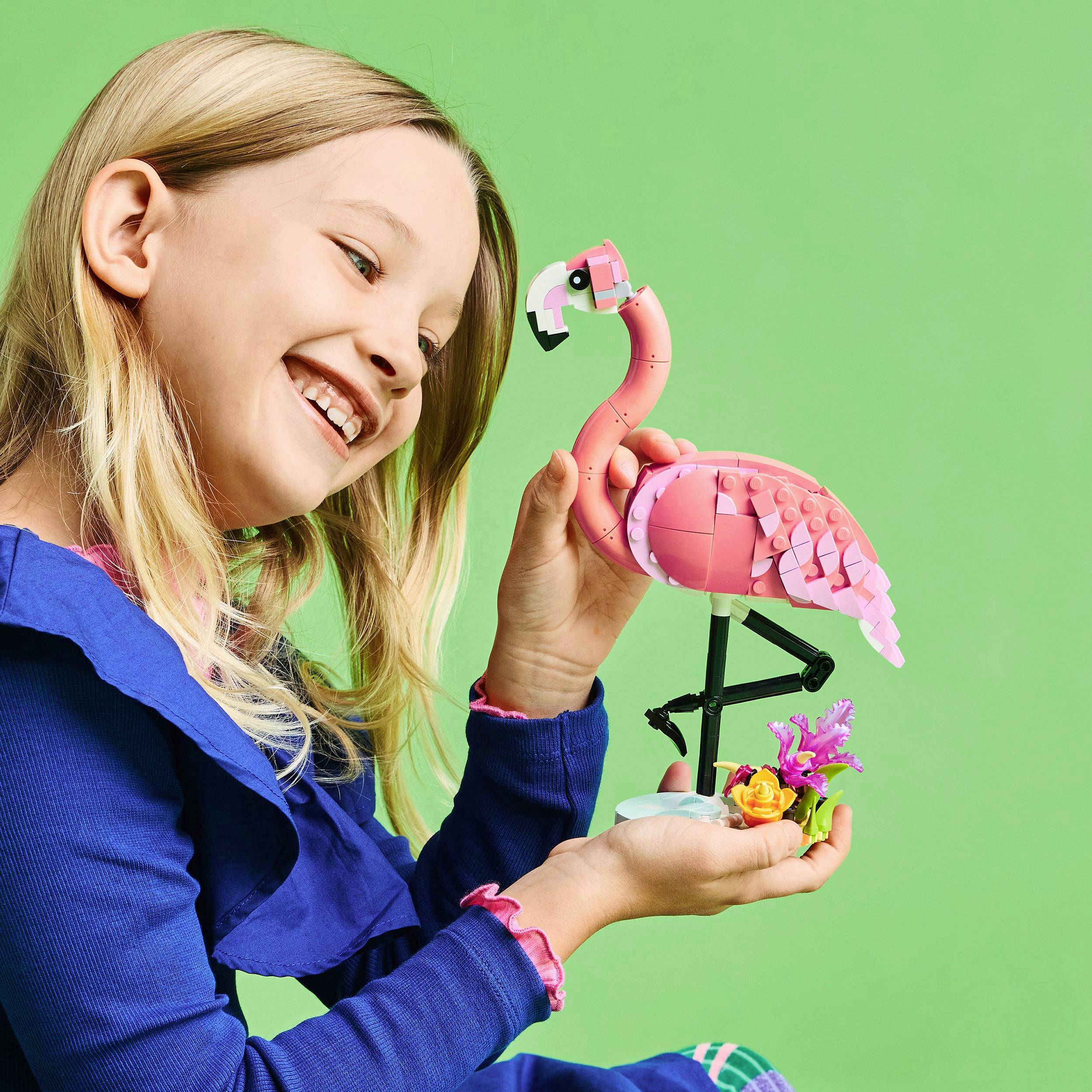 A girl holds a pink Lego flamingo model in front of a green background and smiles.