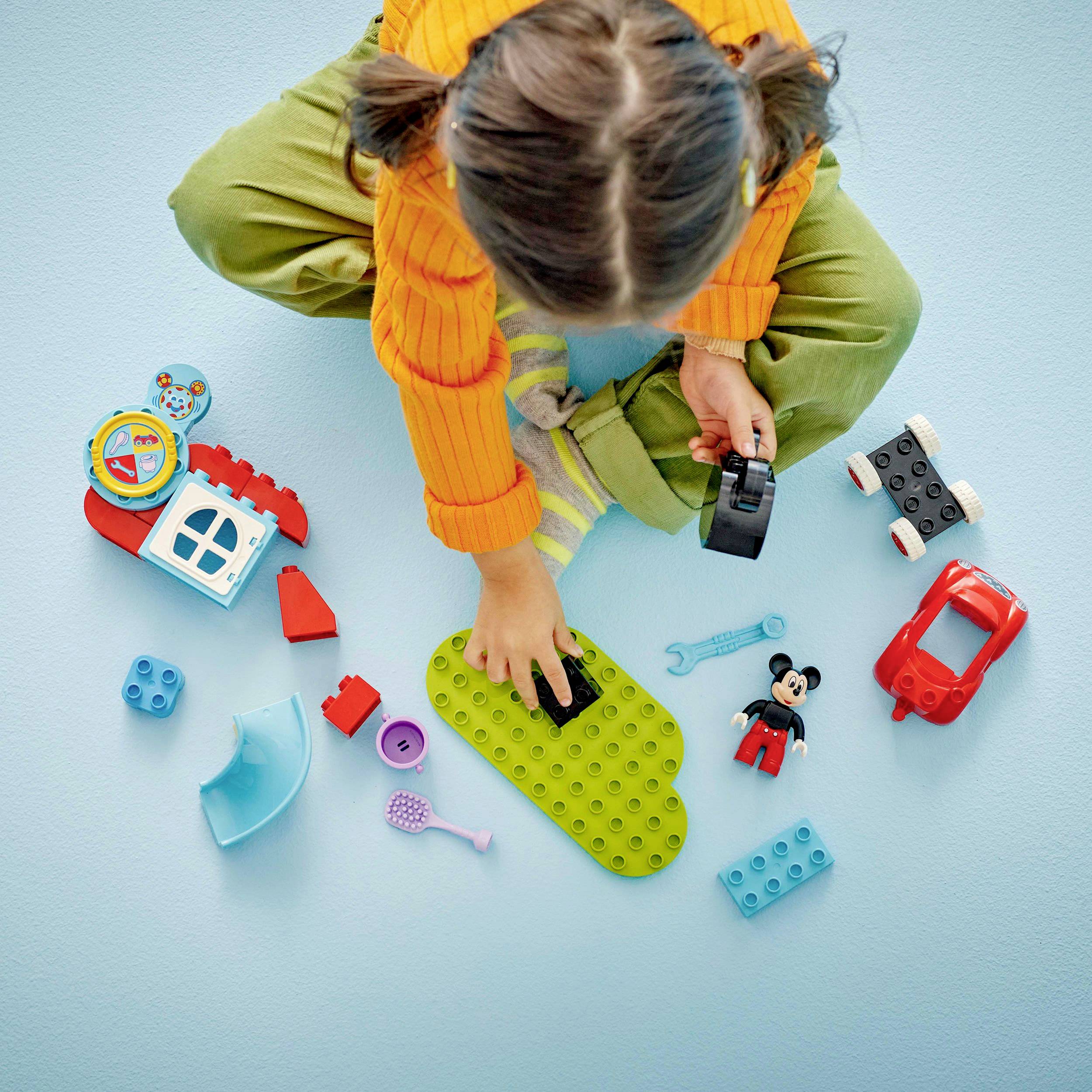 A child in an orange shirt is playing on a light blue floor with colourful building blocks and toys, including a Mickey Mouse figure.