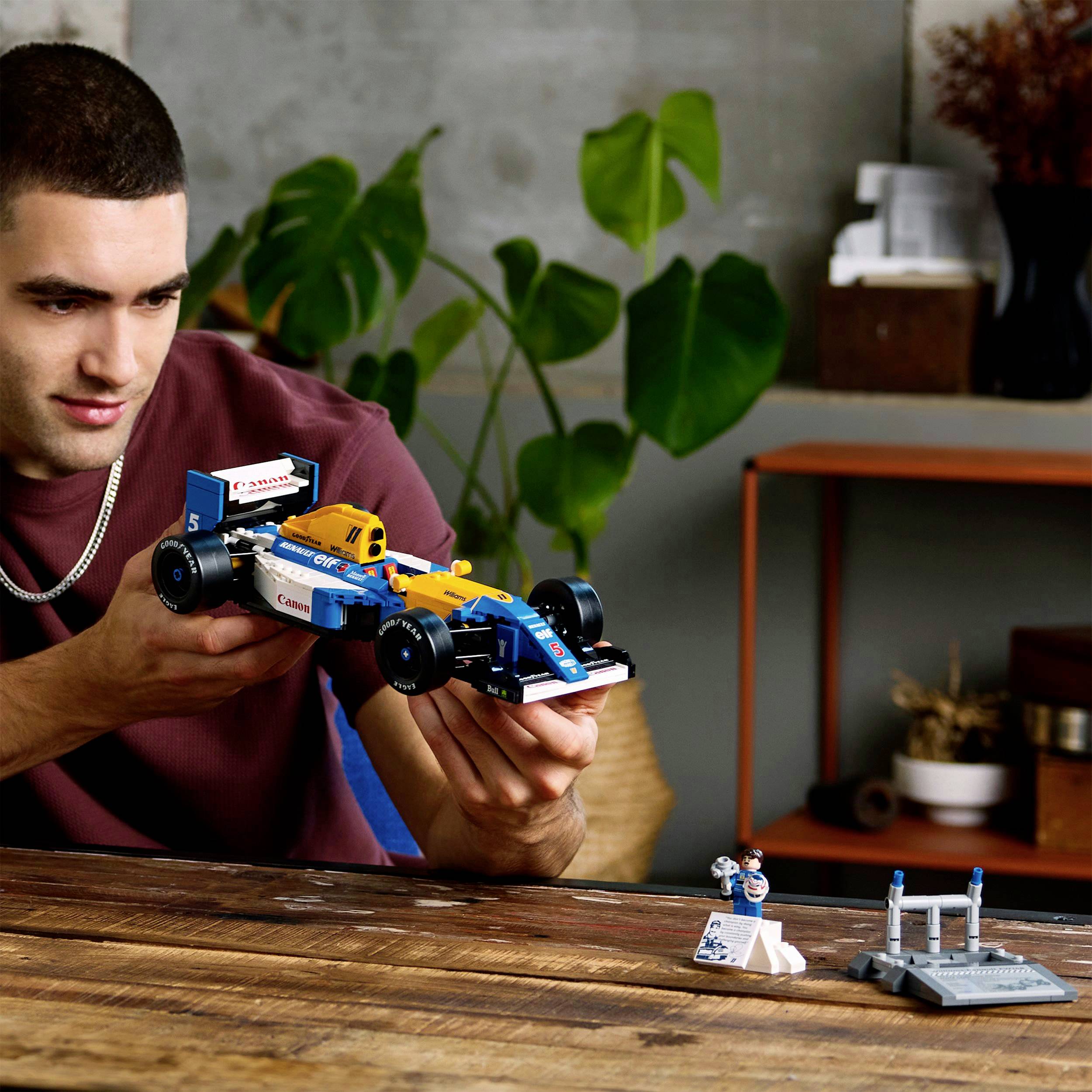 A man is assembling a model car using blue and orange building blocks. Shelves with decorations are visible in the background.