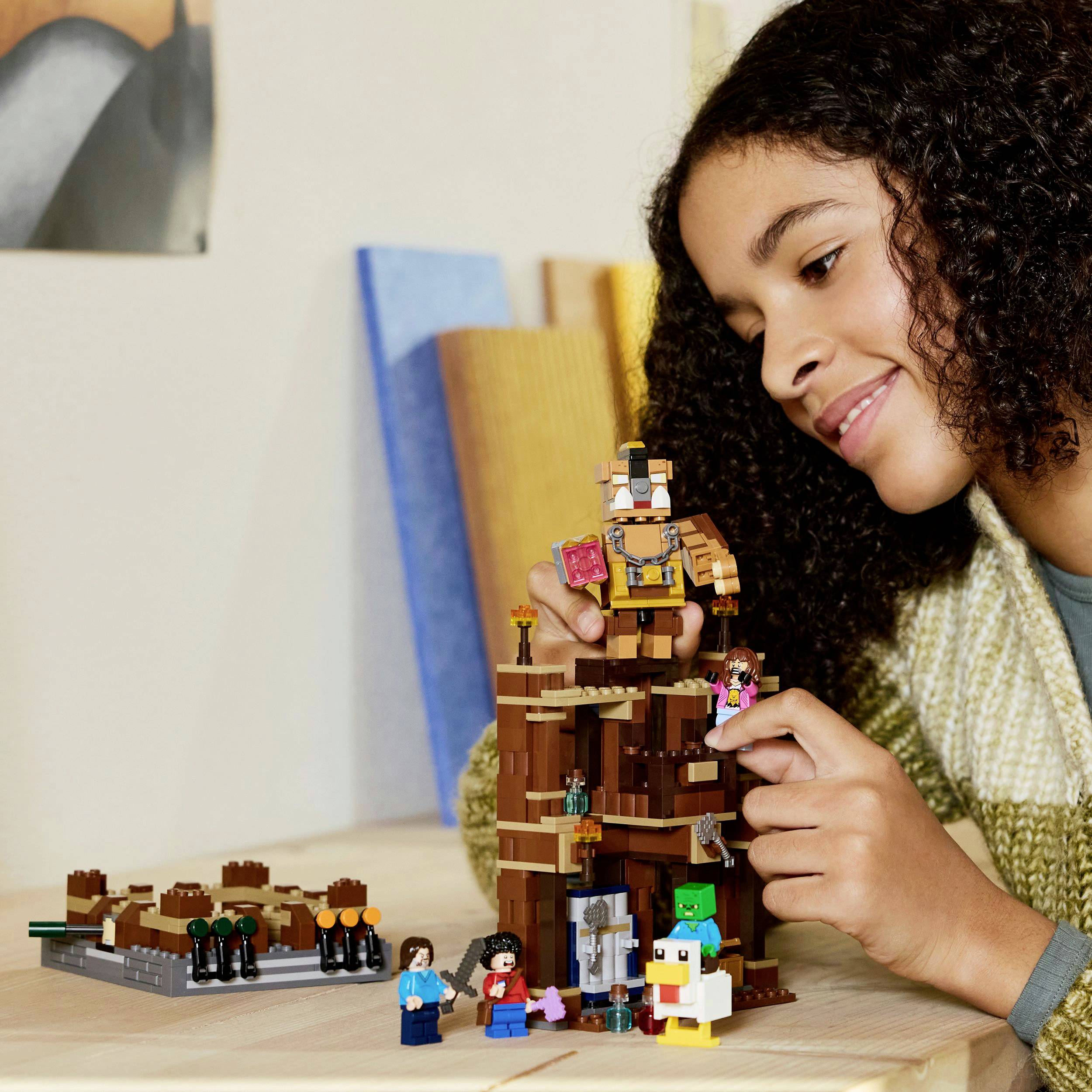 A child is smiling while playing with a Lego set on a table, which includes figures and a structured construction.