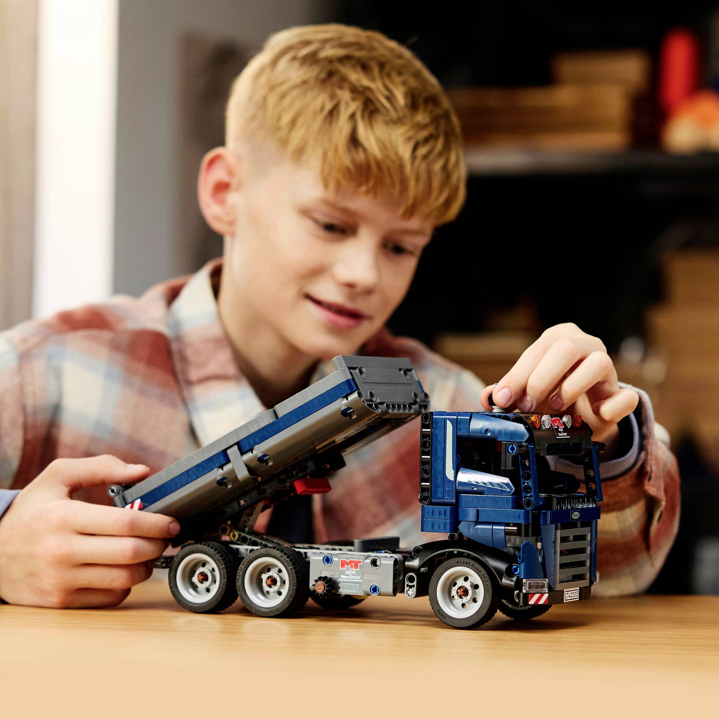 A child is joyfully playing with a blue model lorry, which has a tipping load bed, in a room with a blurred background.