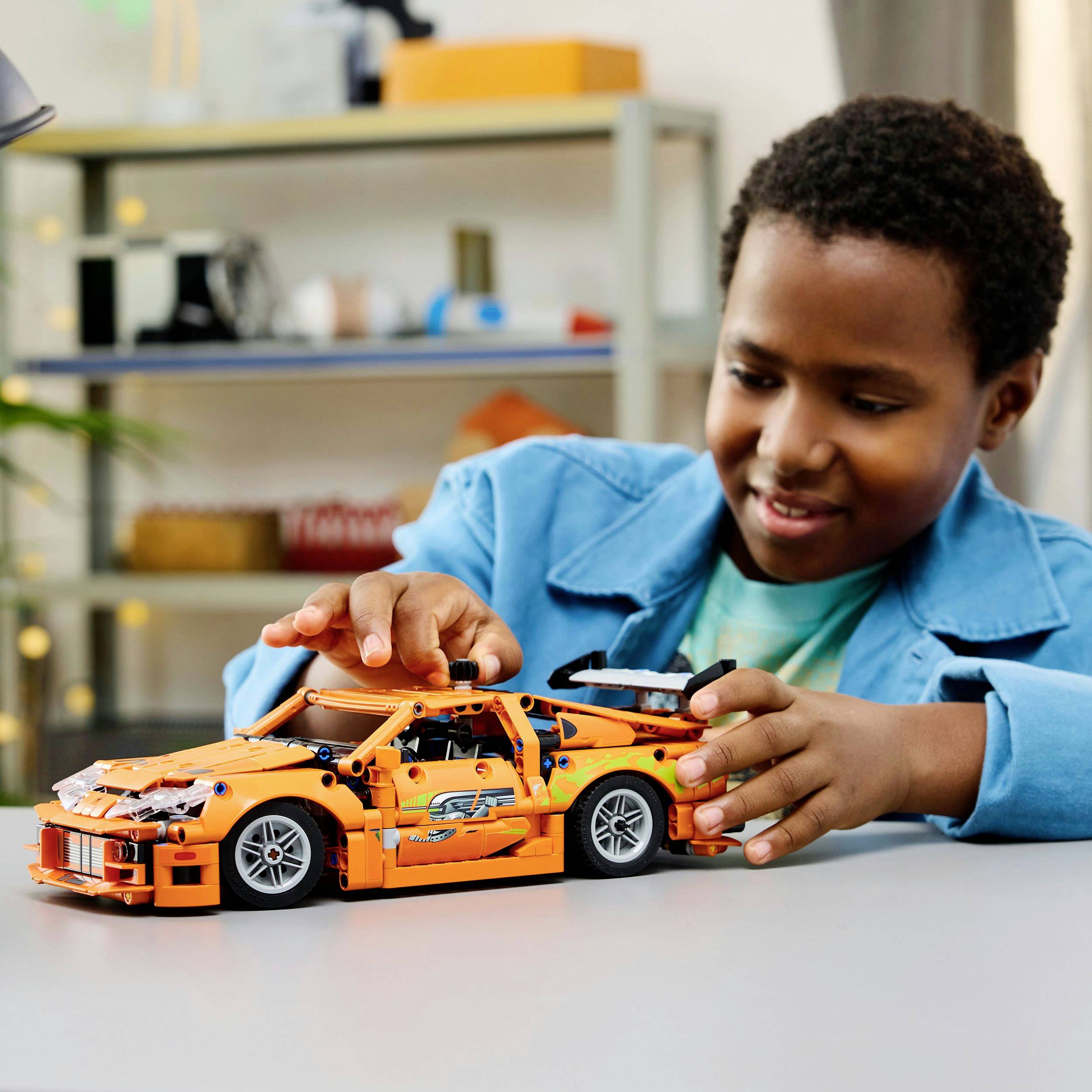 A child plays enthusiastically with an orange toy car on a table, carefully steering it with their hands.