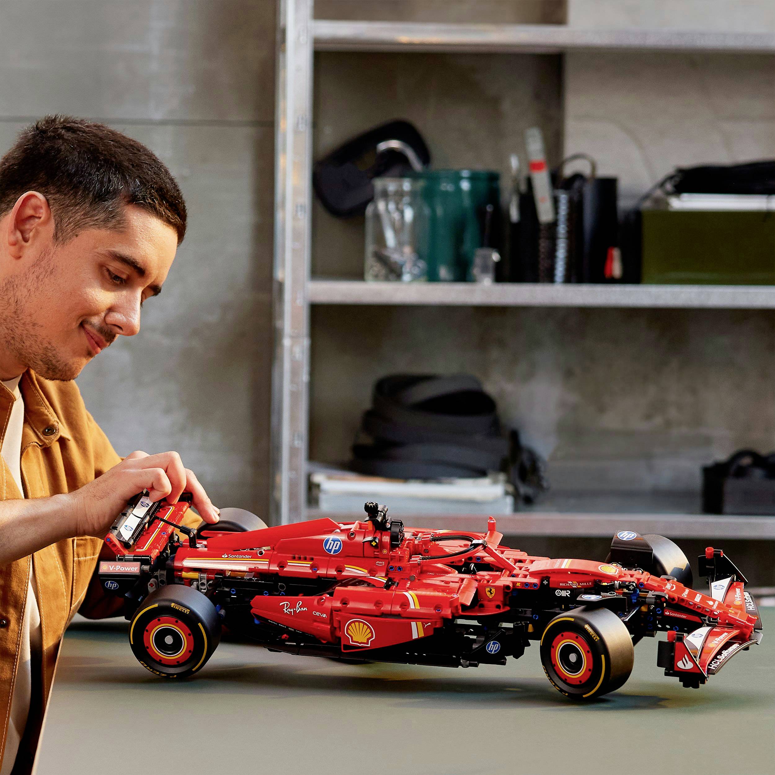 A man is examining a detailed model of a red racing car on a table. In the background, shelves with various objects are visible.