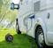 A motorhome is parked on a green meadow and connected to a power source via a cable. Trees and a river can be seen in the background.