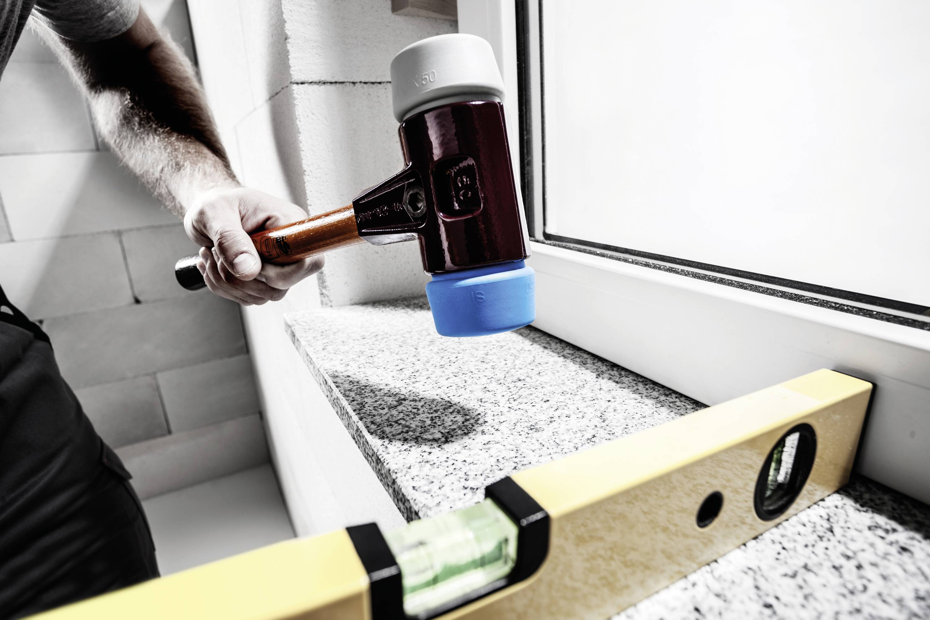 A person is working on a granite slab next to a window frame using a rubber mallet. A spirit level is being used for levelling.
