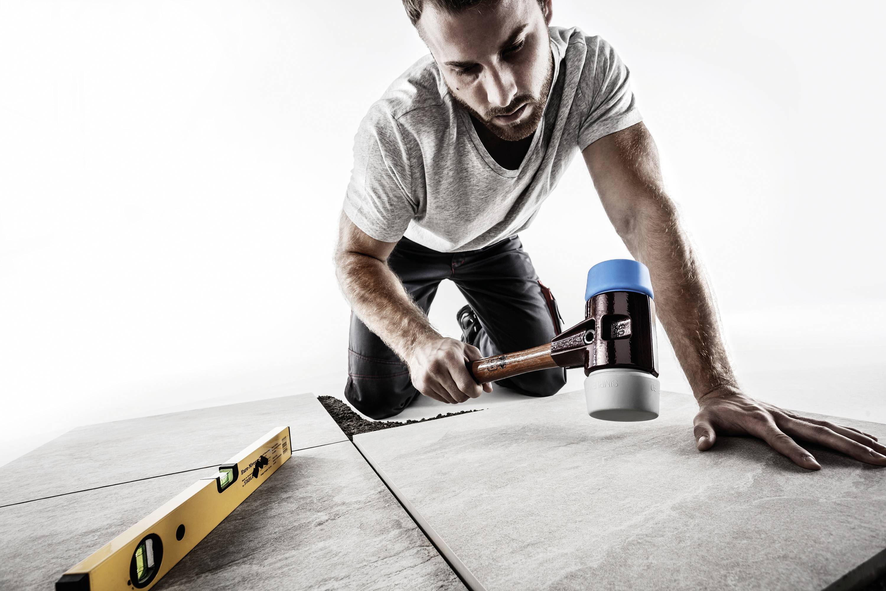 A man is kneeling on the floor and using a rubber mallet to align tiles. A spirit level is lying nearby.