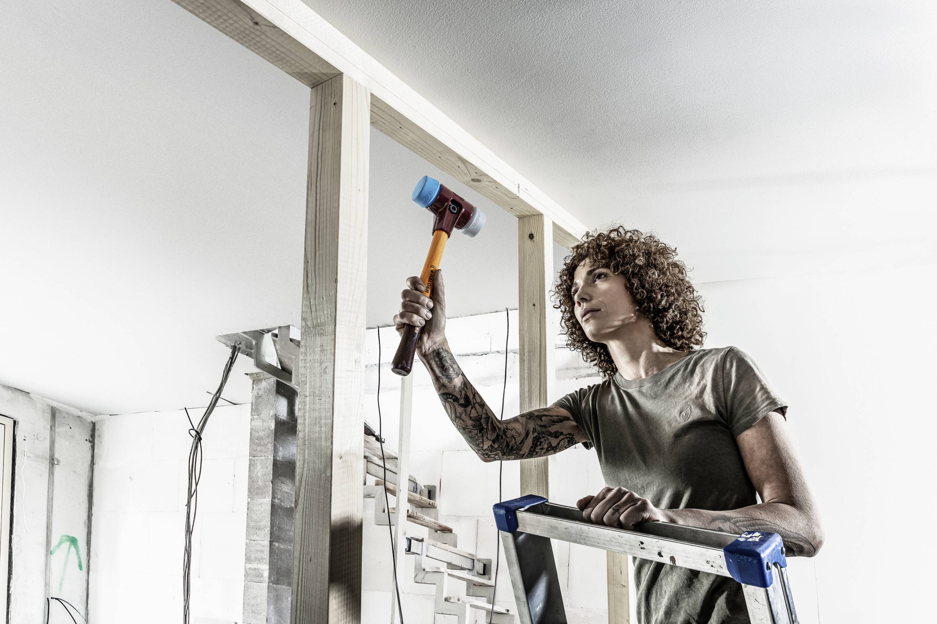 A person on a ladder working on a wooden frame with a hammer. Construction equipment can be seen in the background.
