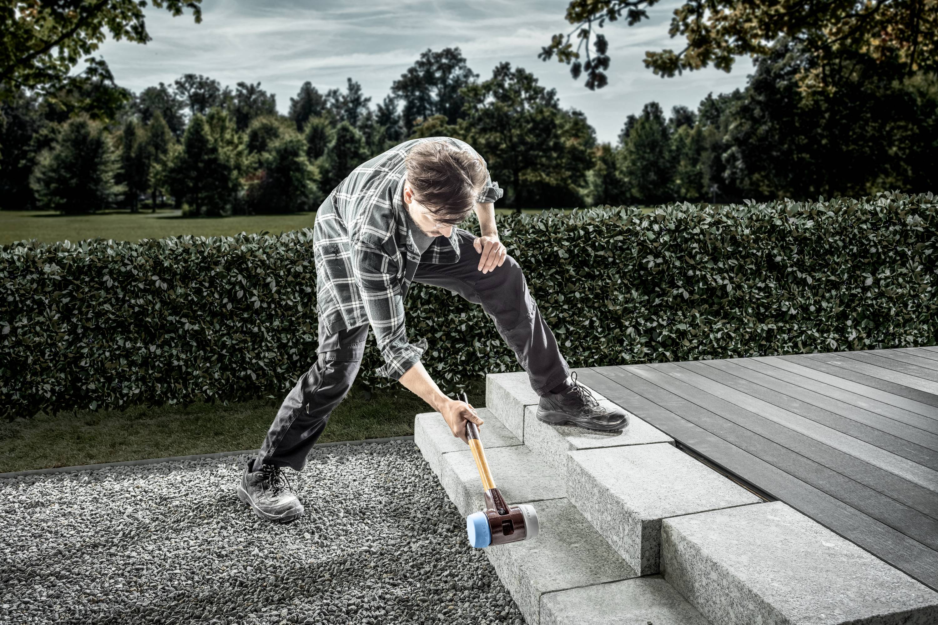 A man is working on stone steps outdoors using a large hammer. A hedge and trees can be seen in the background.