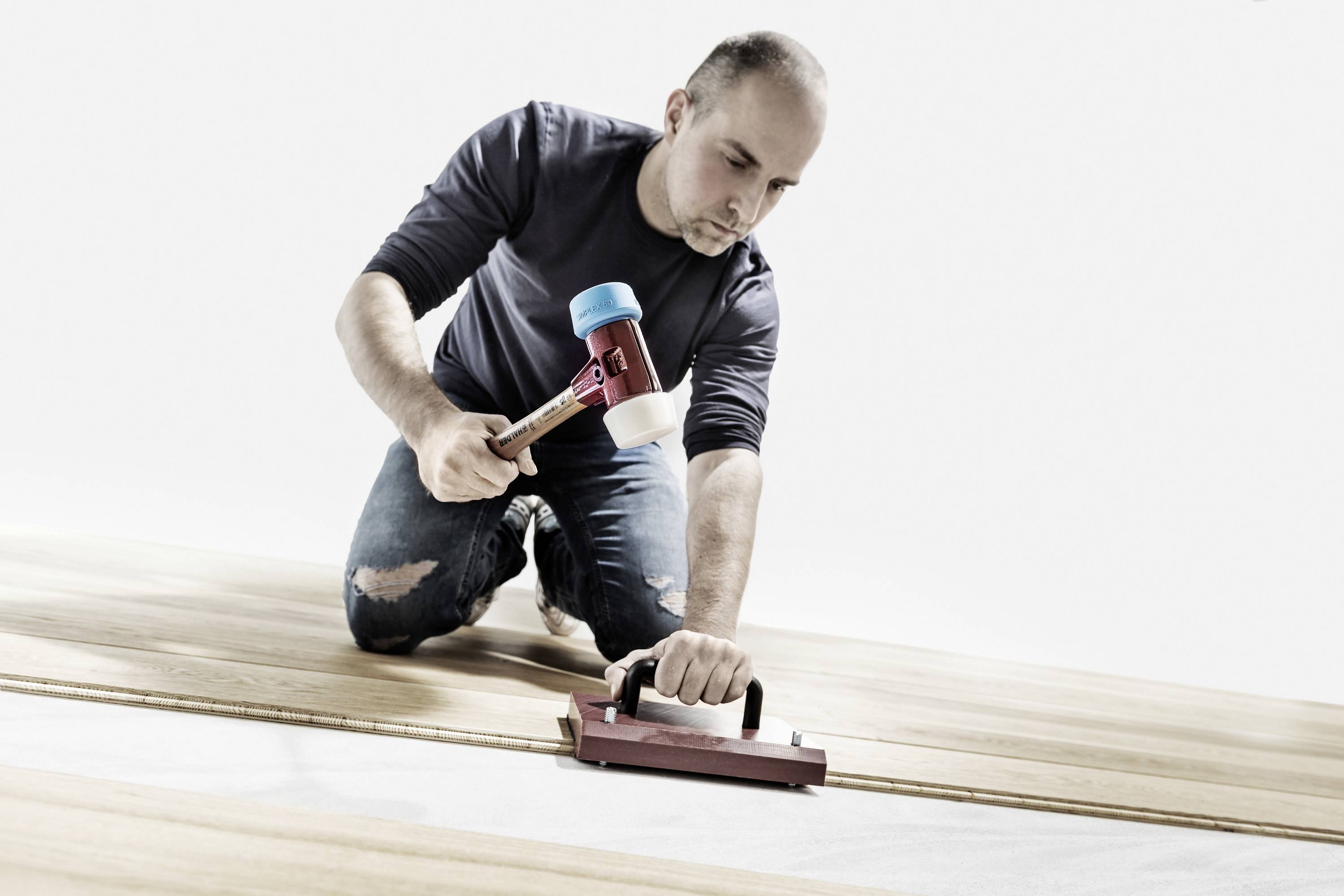 A man is kneeling on a wooden floor and using a tool to lay the floorboards. He is holding a hammer in his other hand.