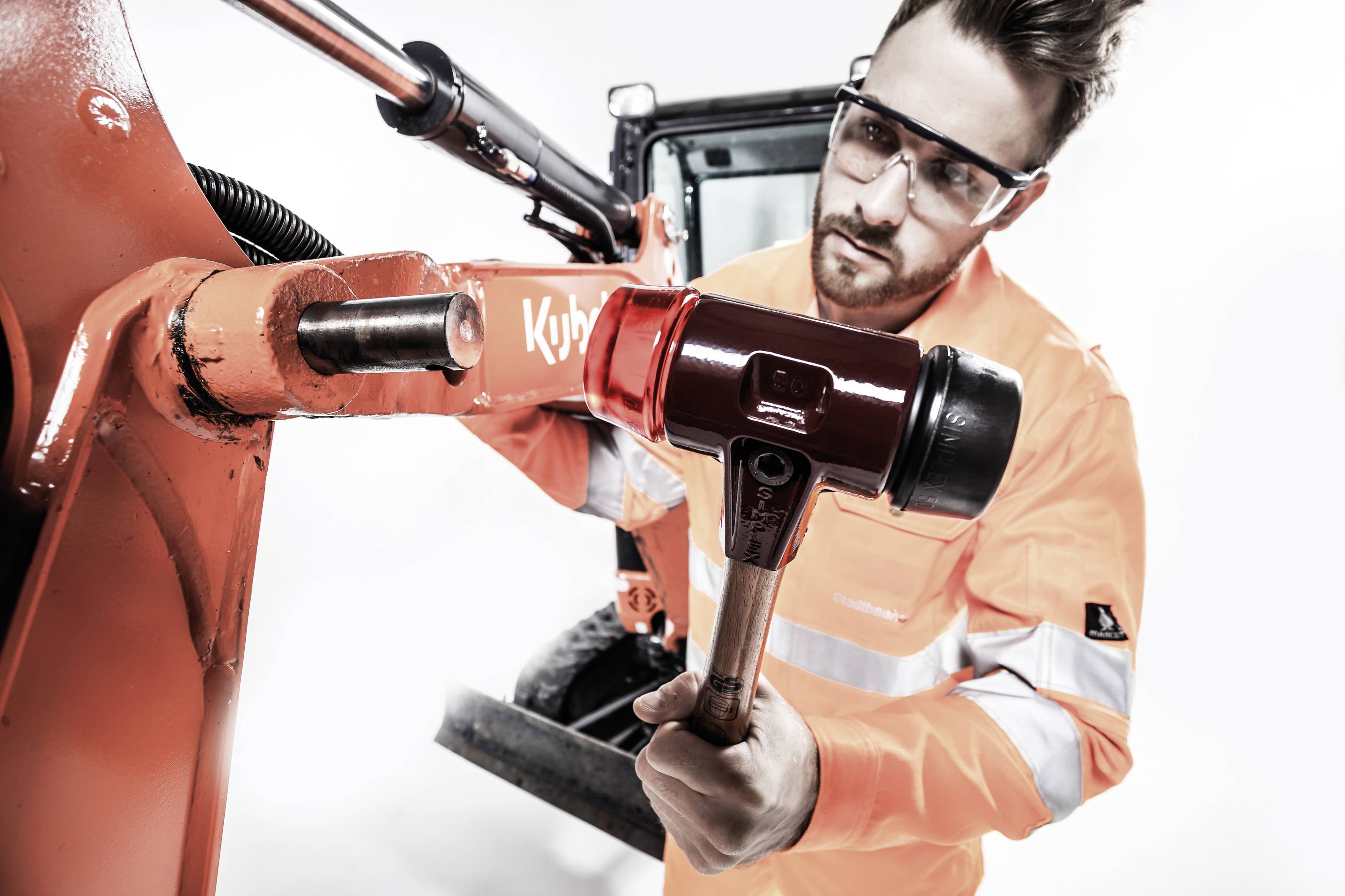A man wearing safety glasses and high-visibility orange protective clothing is working with a hammer on a large red machine.