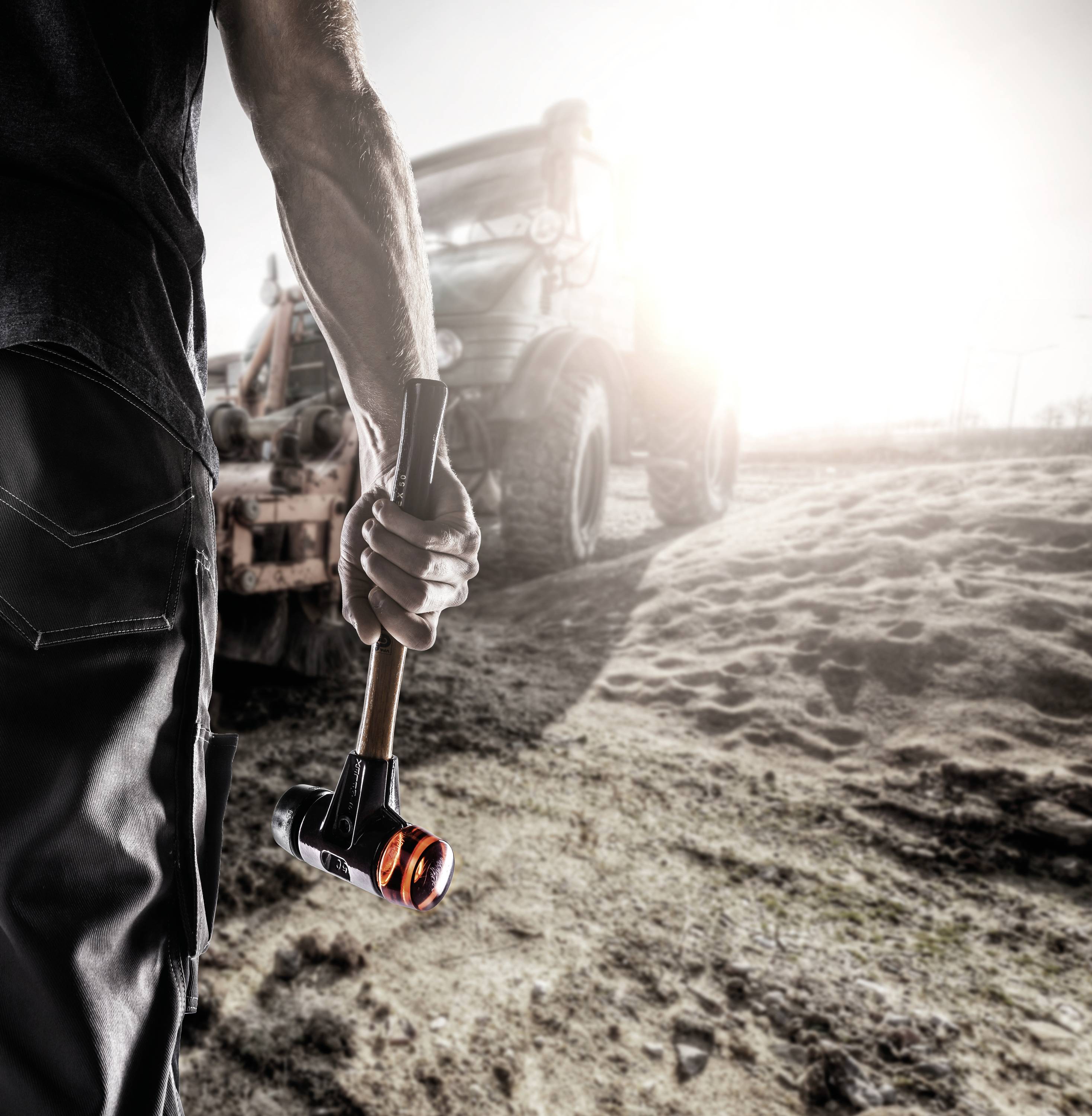 A construction worker stands in the foreground holding a heavy hammer. A blurred tractor is visible in the background.