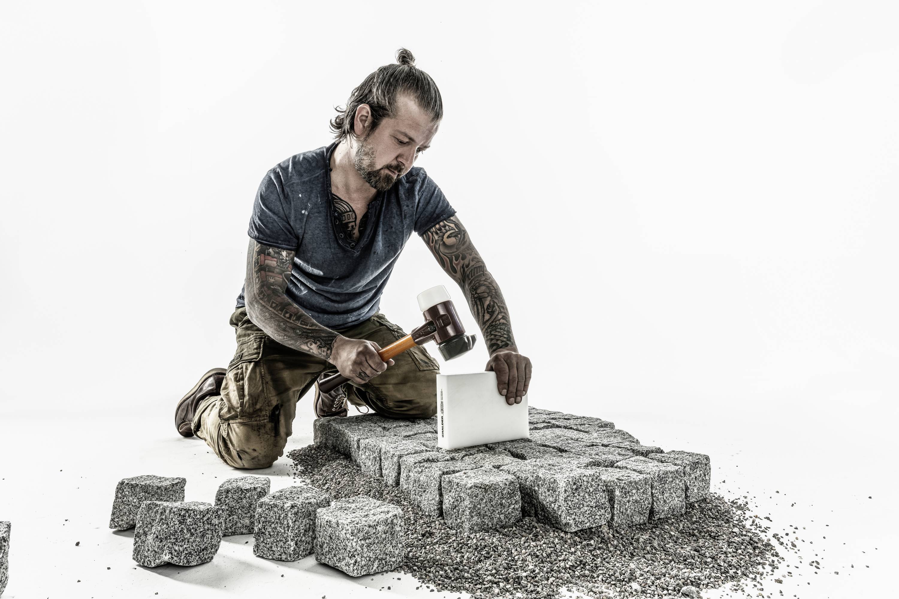 A man is kneeling on the ground and shaping paving stones with a hammer and a moulding block. Around him are already formed stones.