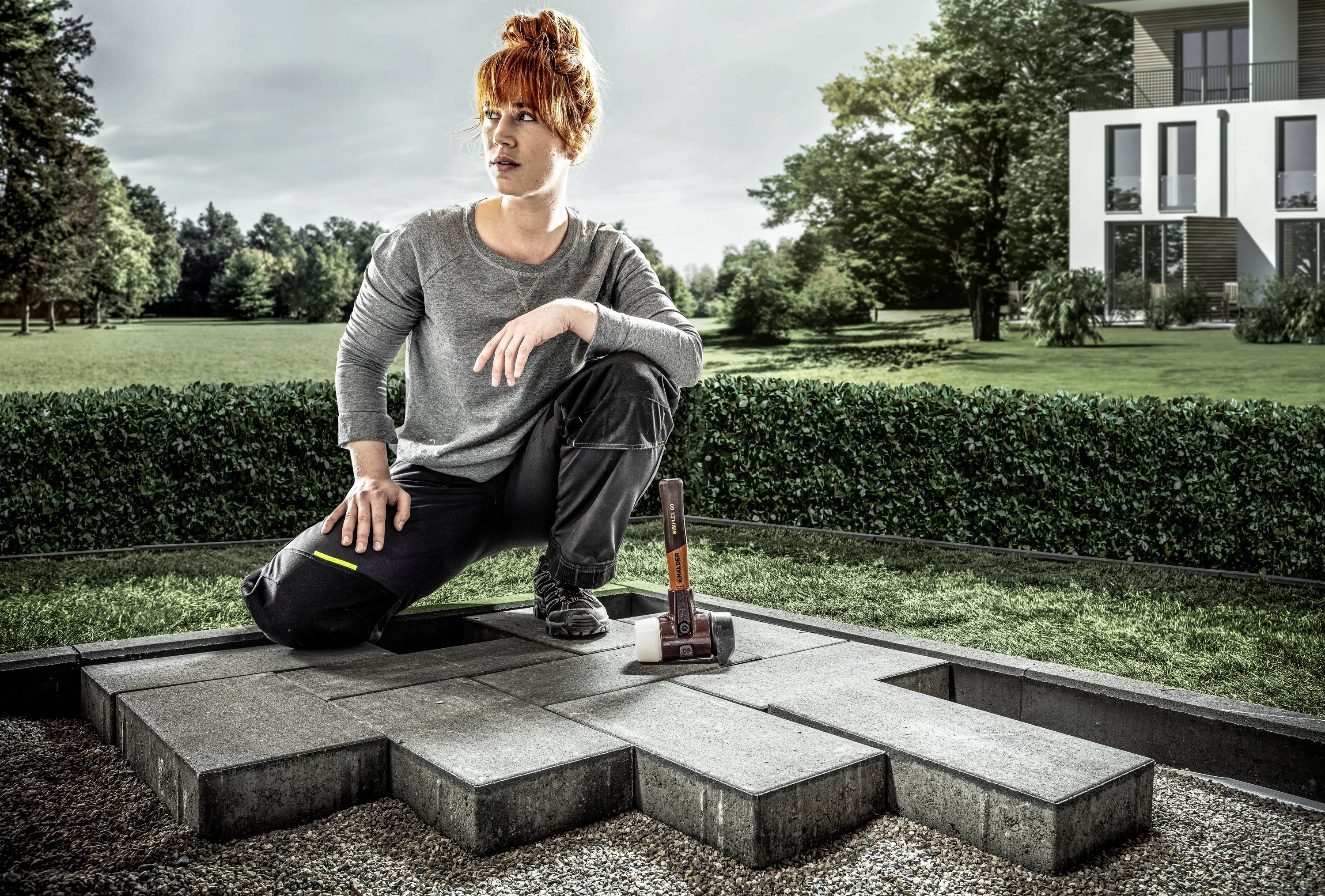 A person is kneeling on paved paving stones in a garden and holding a hammer. Hedges and a house can be seen in the background.
