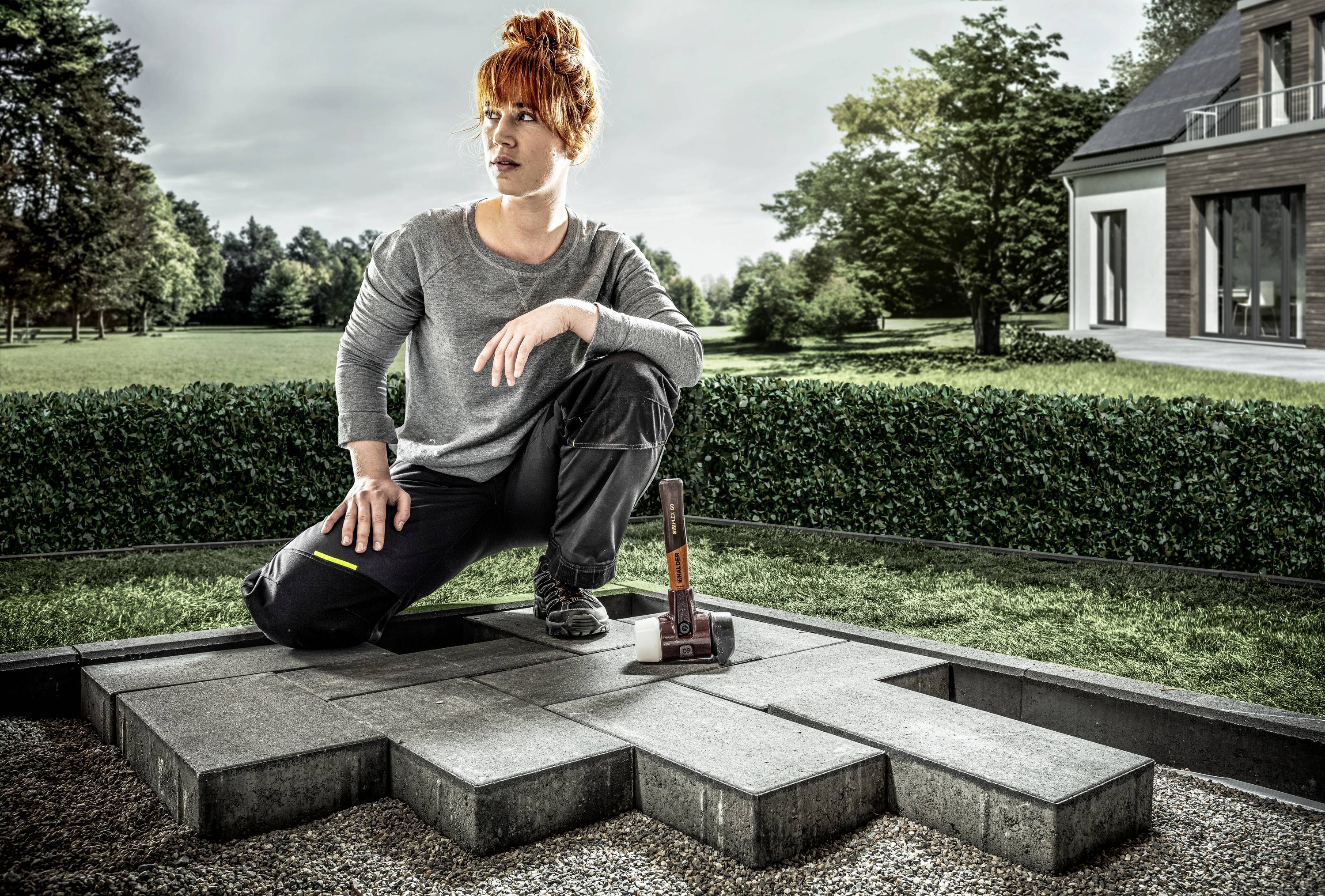A person in work attire is kneeling on paved stones in a garden, with a hammer beside them, surrounded by lawn and a modern house.