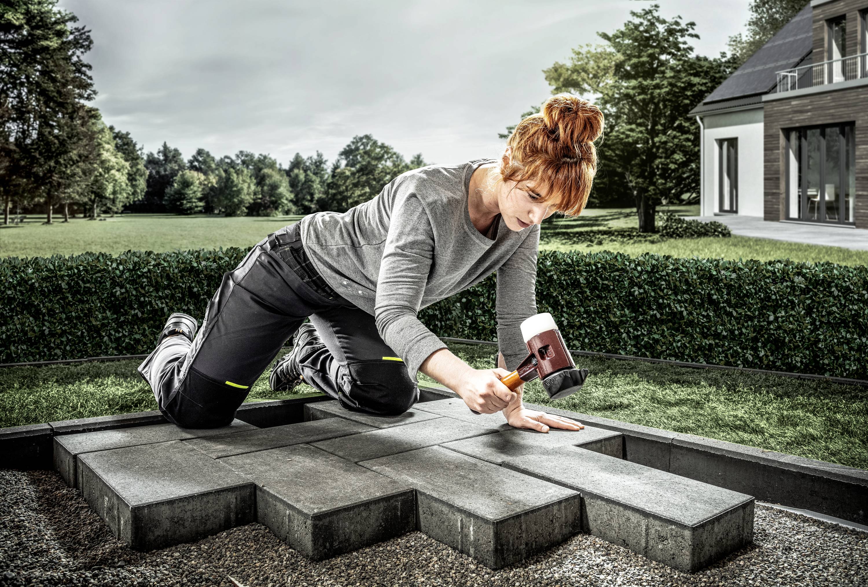 A woman is kneeling on a patio and laying dark grey paving stones with a rubber mallet. A garden and a modern house can be seen in the background.