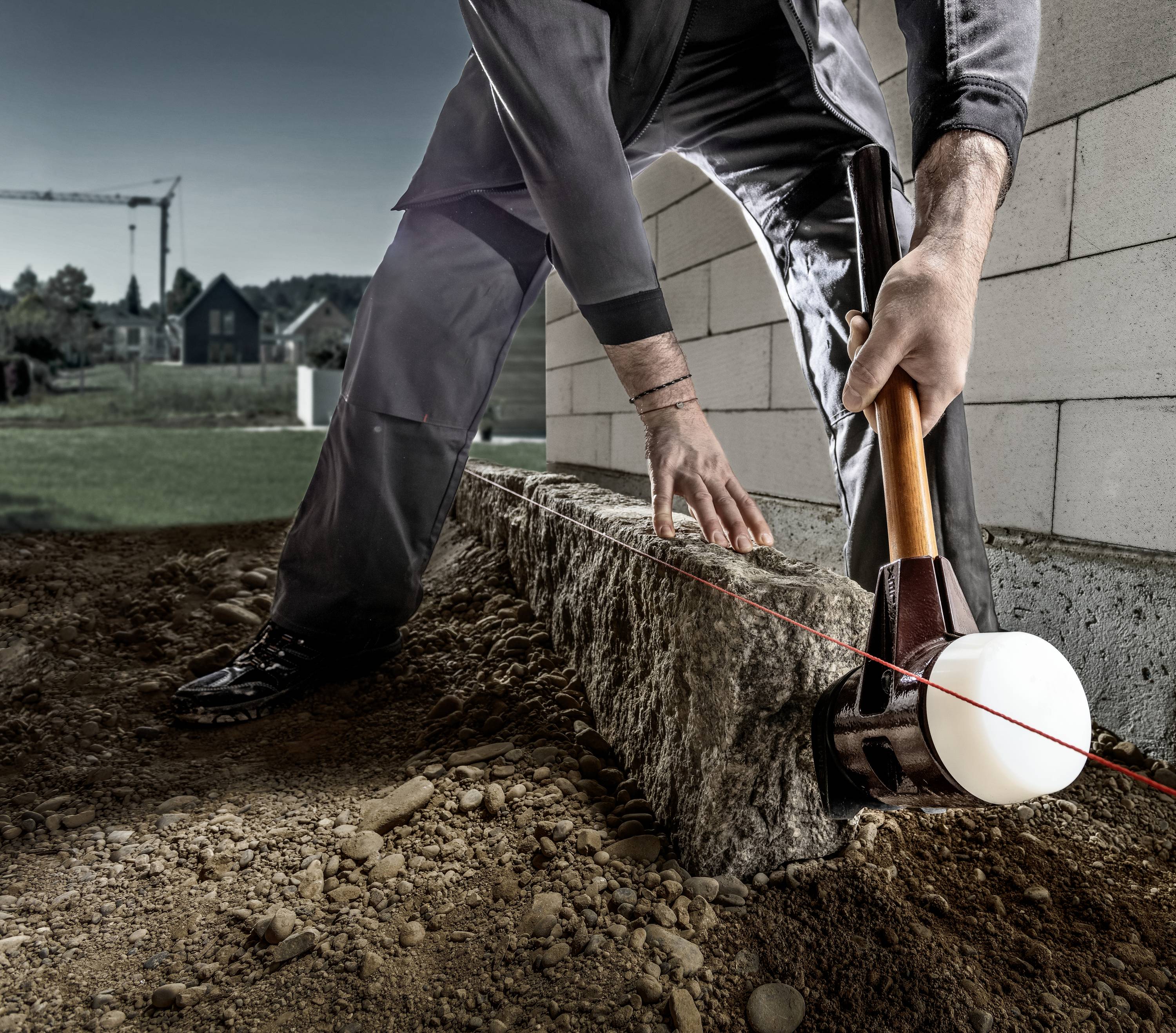 A person is aligning a stone with a hammer in a row of stones. Construction cranes and a house are visible in the background.