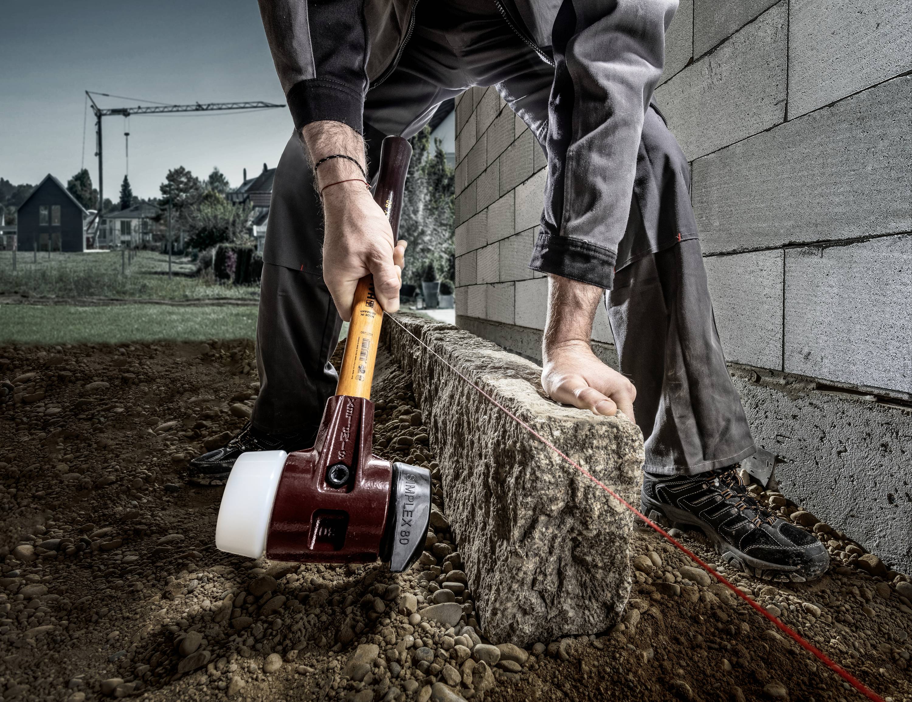 A construction worker positions a concrete block along a red guideline using a large hammer on a building site.