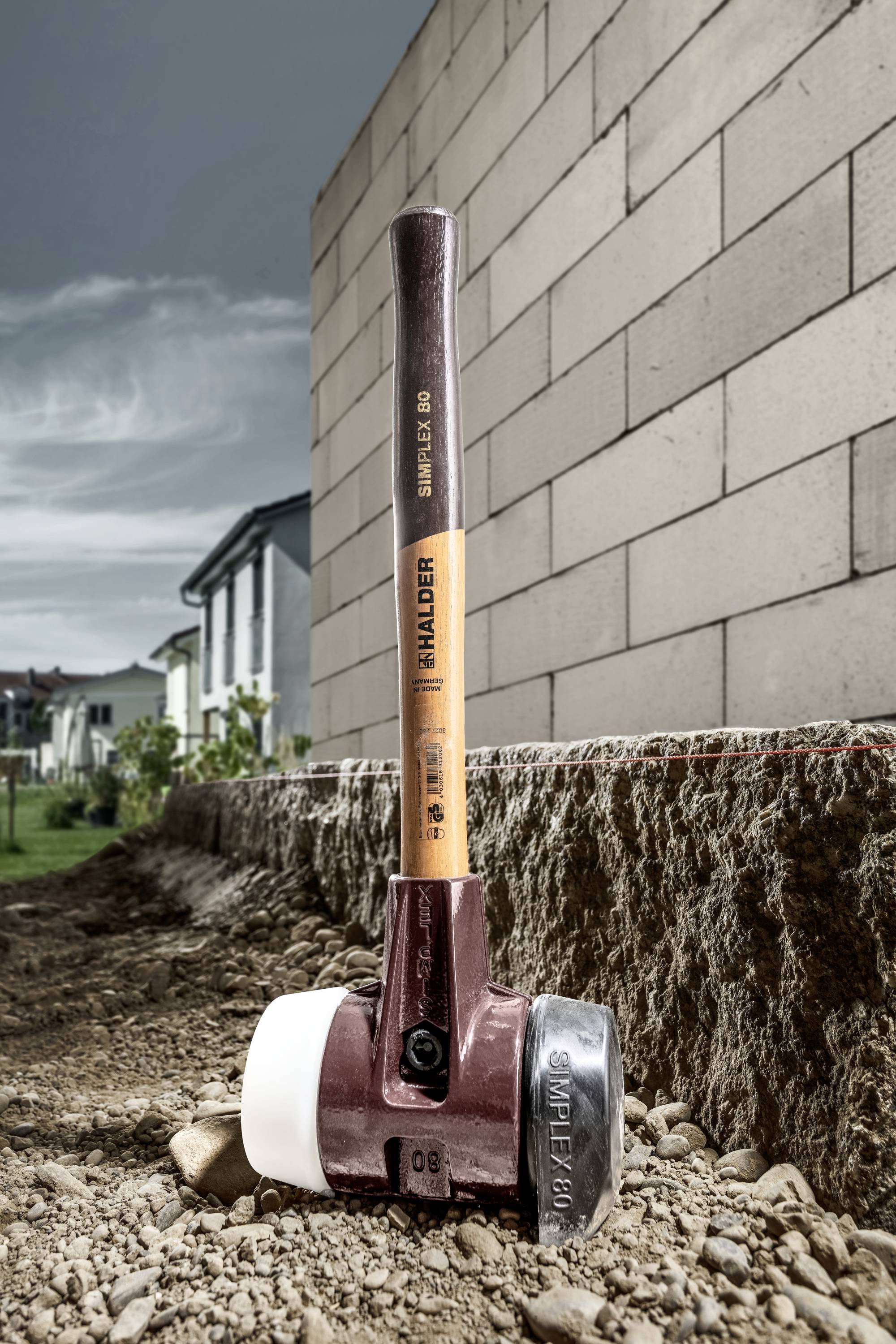 A large construction tool, similar to a hammer, stands on a building site next to a brick wall. Houses are visible in the background.
