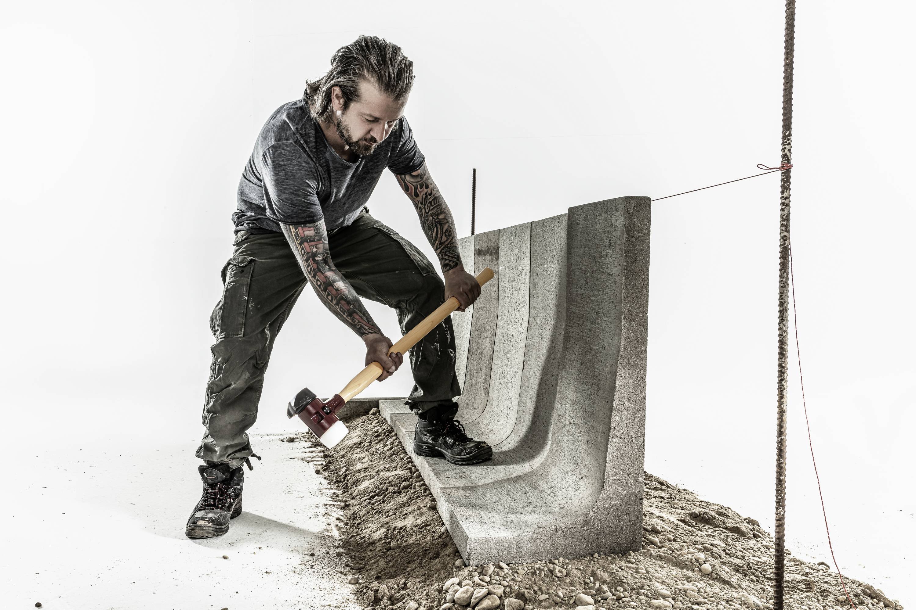 A man is striking a concrete block anchored diagonally in the ground with a sledgehammer. He is wearing work attire.