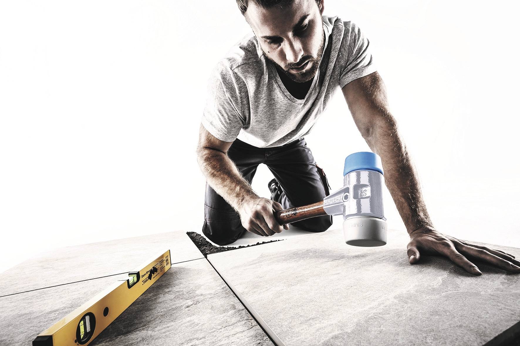 A man is kneeling on the floor and laying tiles. In one hand, he is holding a rubber mallet, and a spirit level is lying nearby.