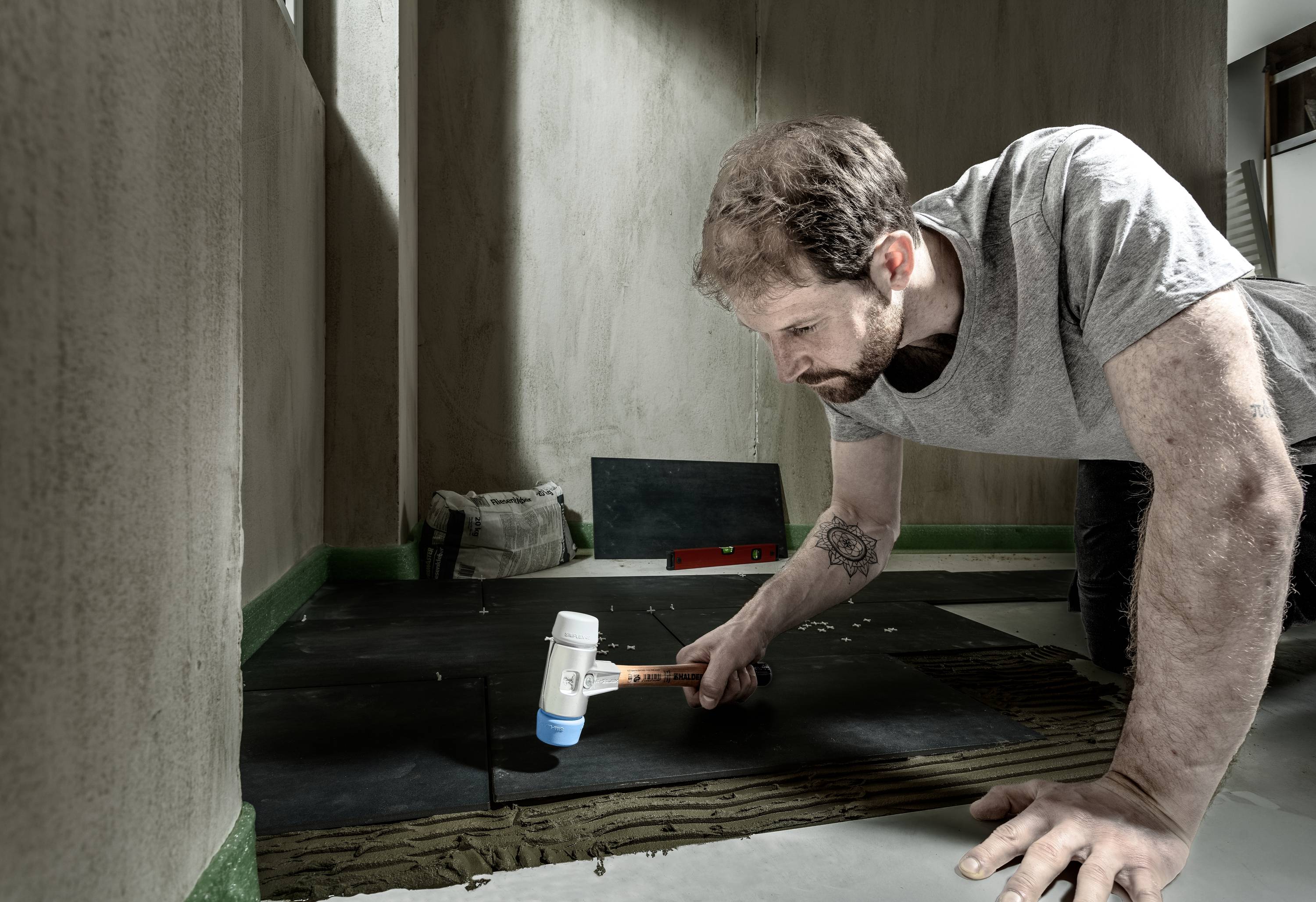 A man is kneeling on the floor and using a specialised tool to lay floor tiles. The environment is bare and unfinished.
