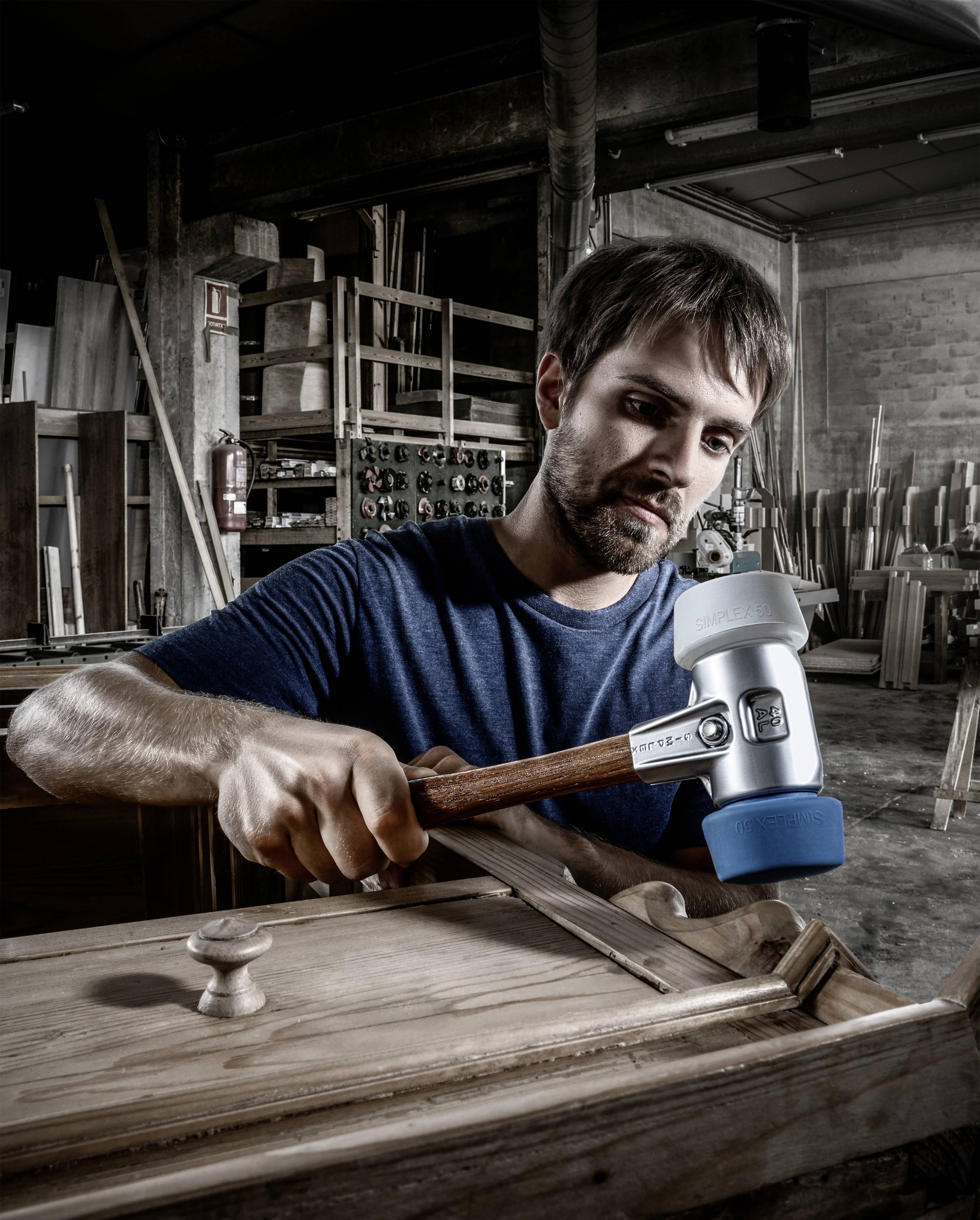 A man is working attentively with a wooden hammer on a piece of furniture in a workshop. Tools and wood are scattered around him.