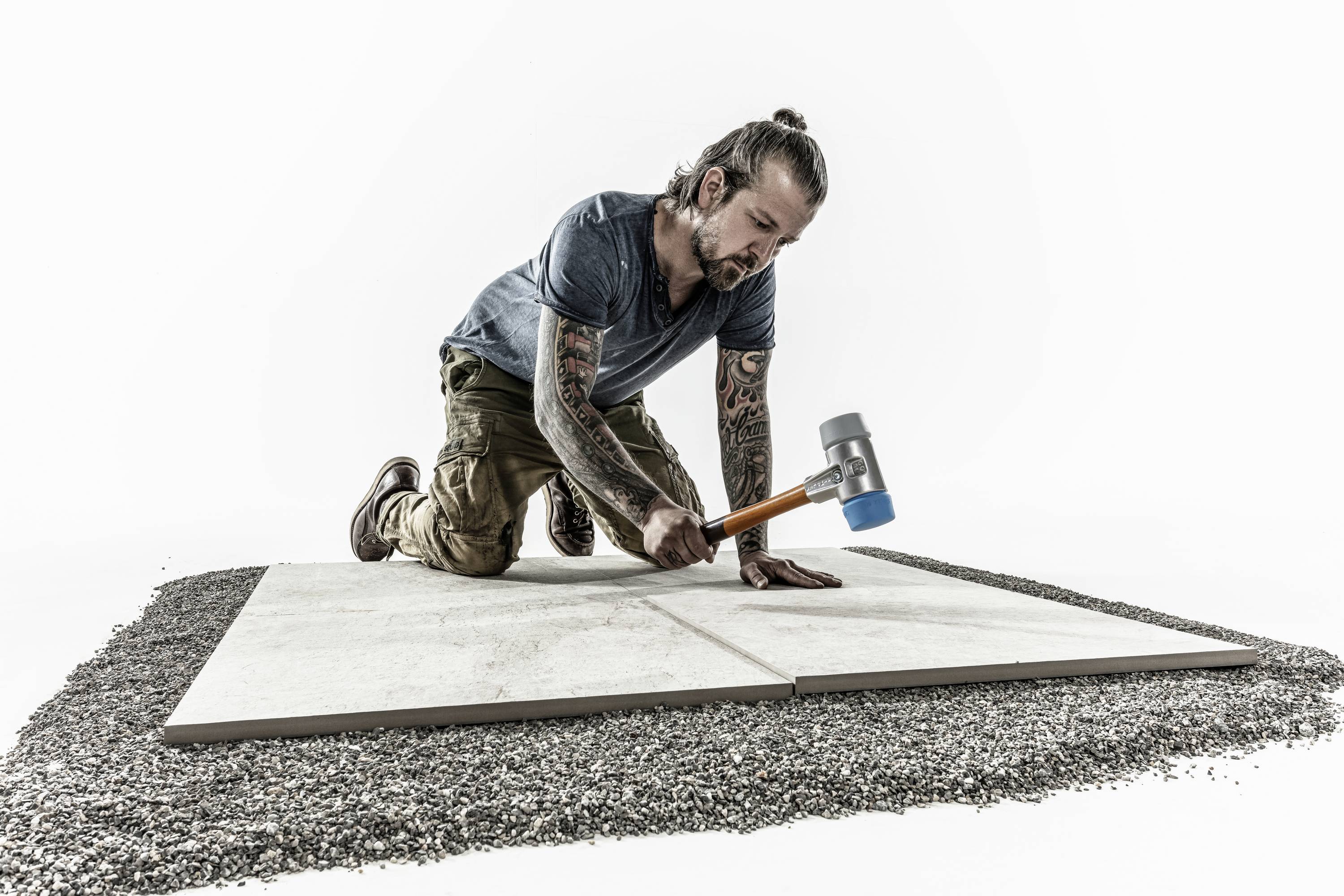 A man is kneeling on gravel and laying large tiles, tapping them into place with a hammer.