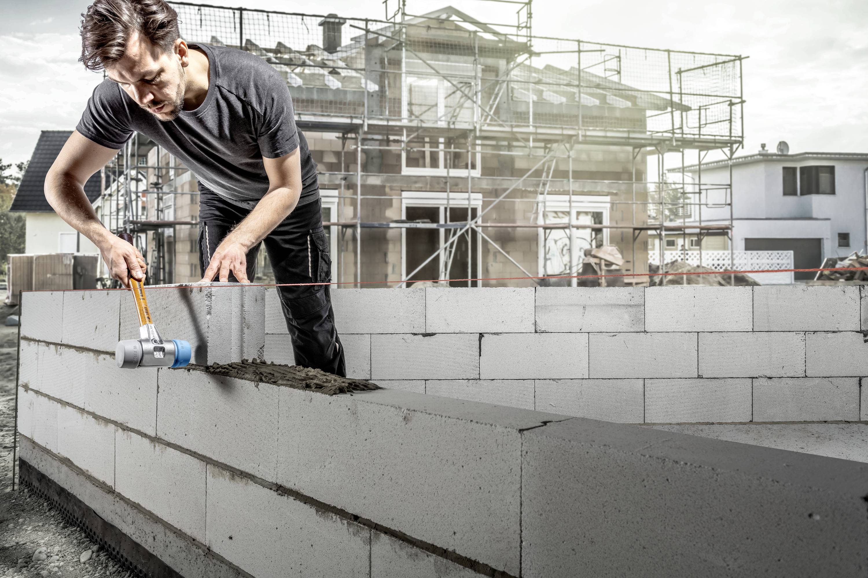 A construction worker is applying cement between concrete blocks on a building site. A scaffolding is visible on a house in the background.