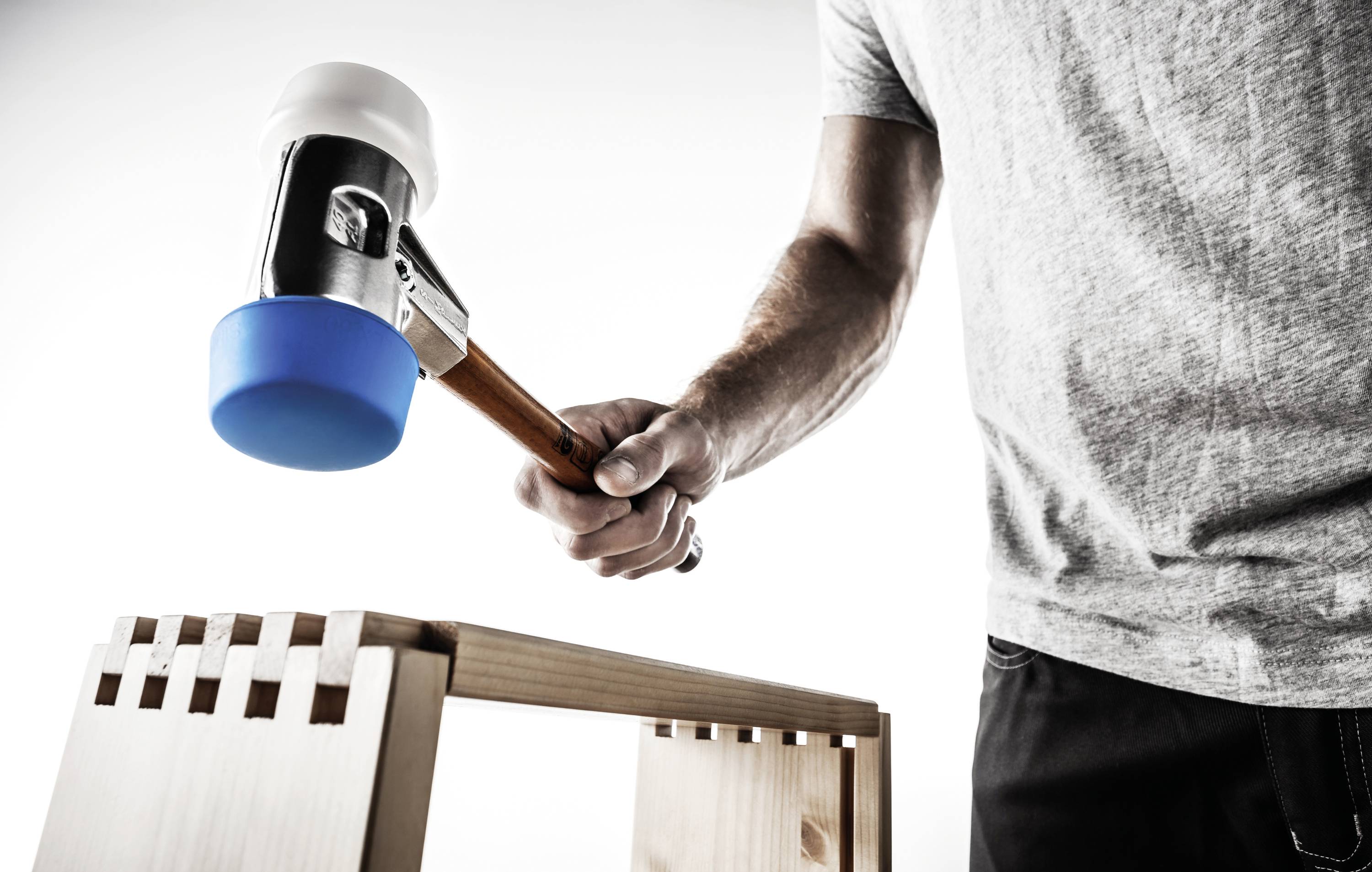 A person is hammering a blue rubber-headed hammer onto a wooden joint. The focus is on craftsmanship and precision.