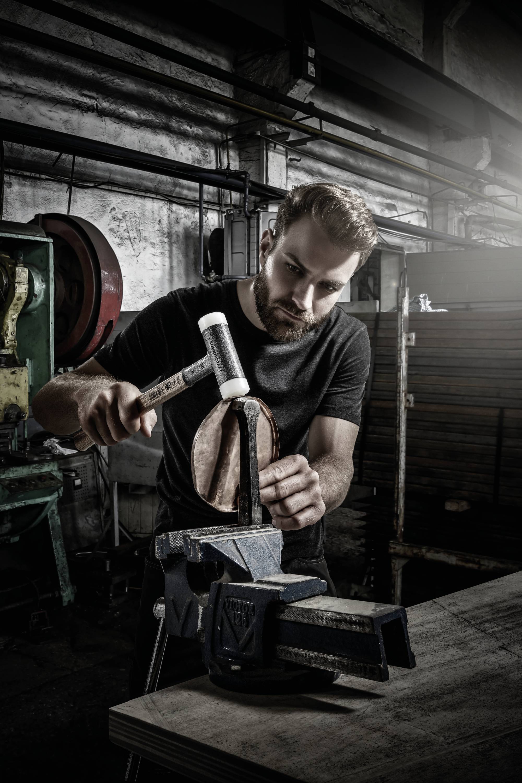 A man is shaping a copper bowl with a hammer in a workshop. Tools and materials can be seen everywhere.