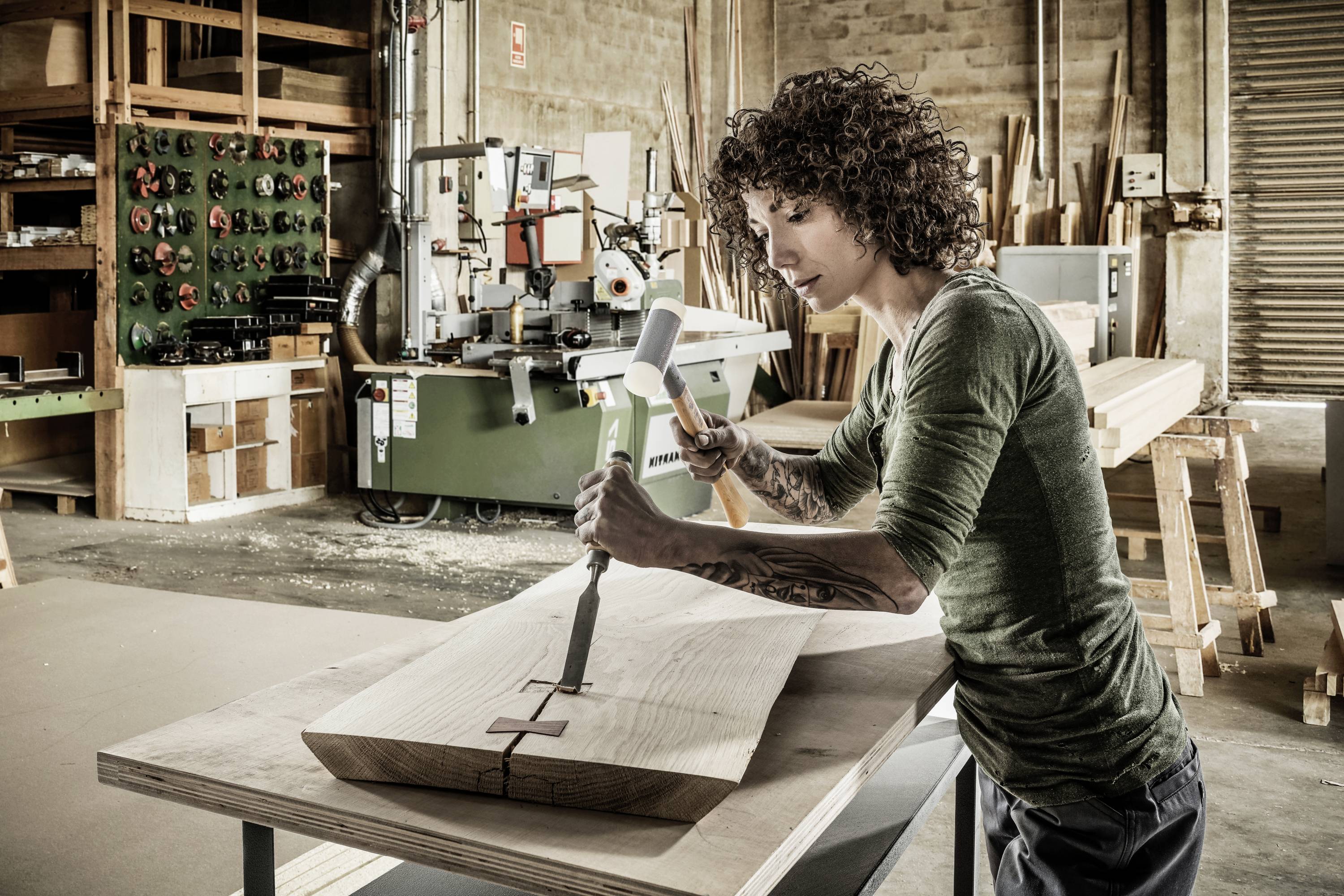 A person is working in a workshop and using a chisel and hammer to craft wood. Machines are visible in the background.