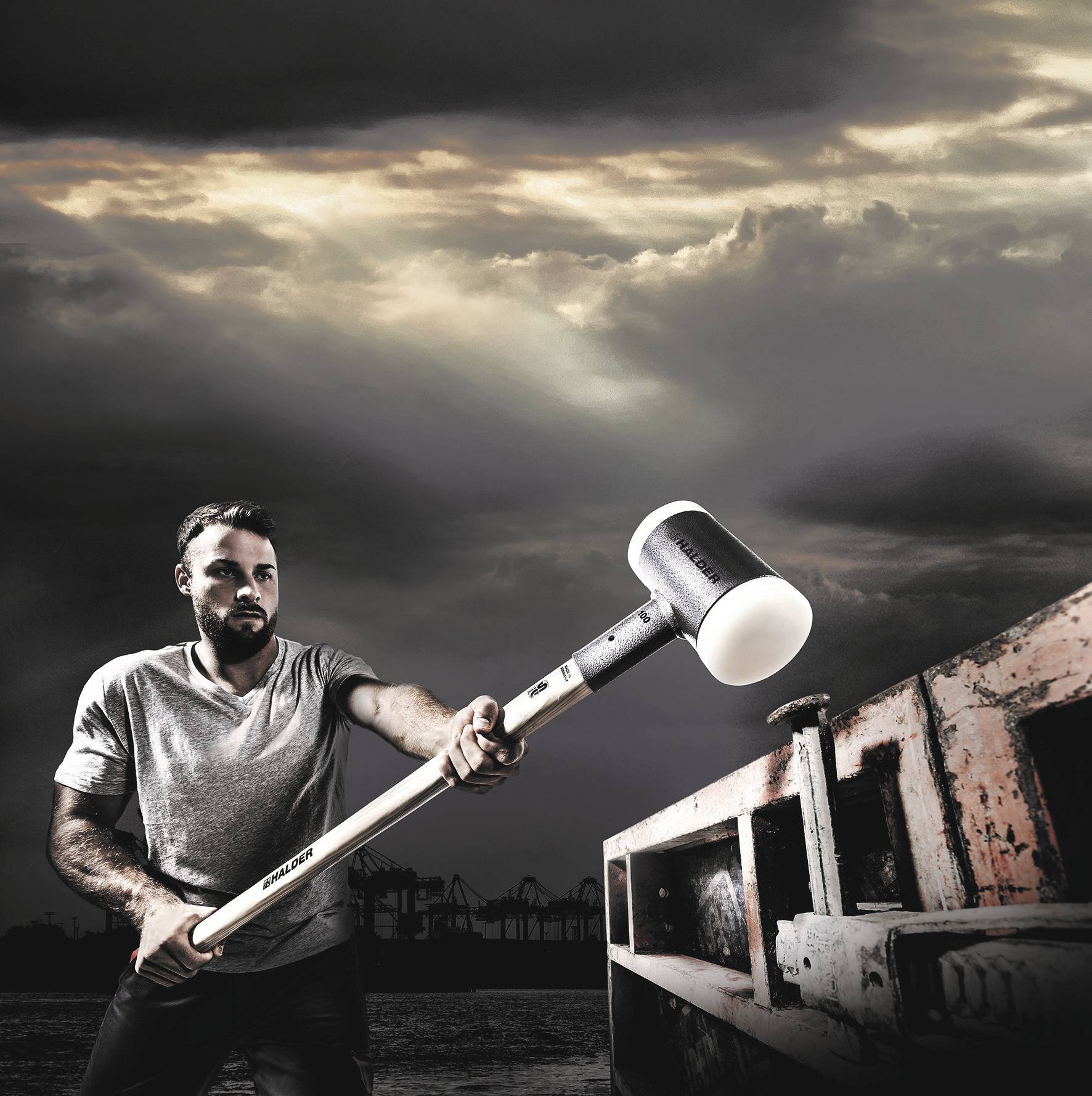 Man in a T-shirt striking a shipping container with a large hammer in an industrial harbour, with a dramatic sky in the background.