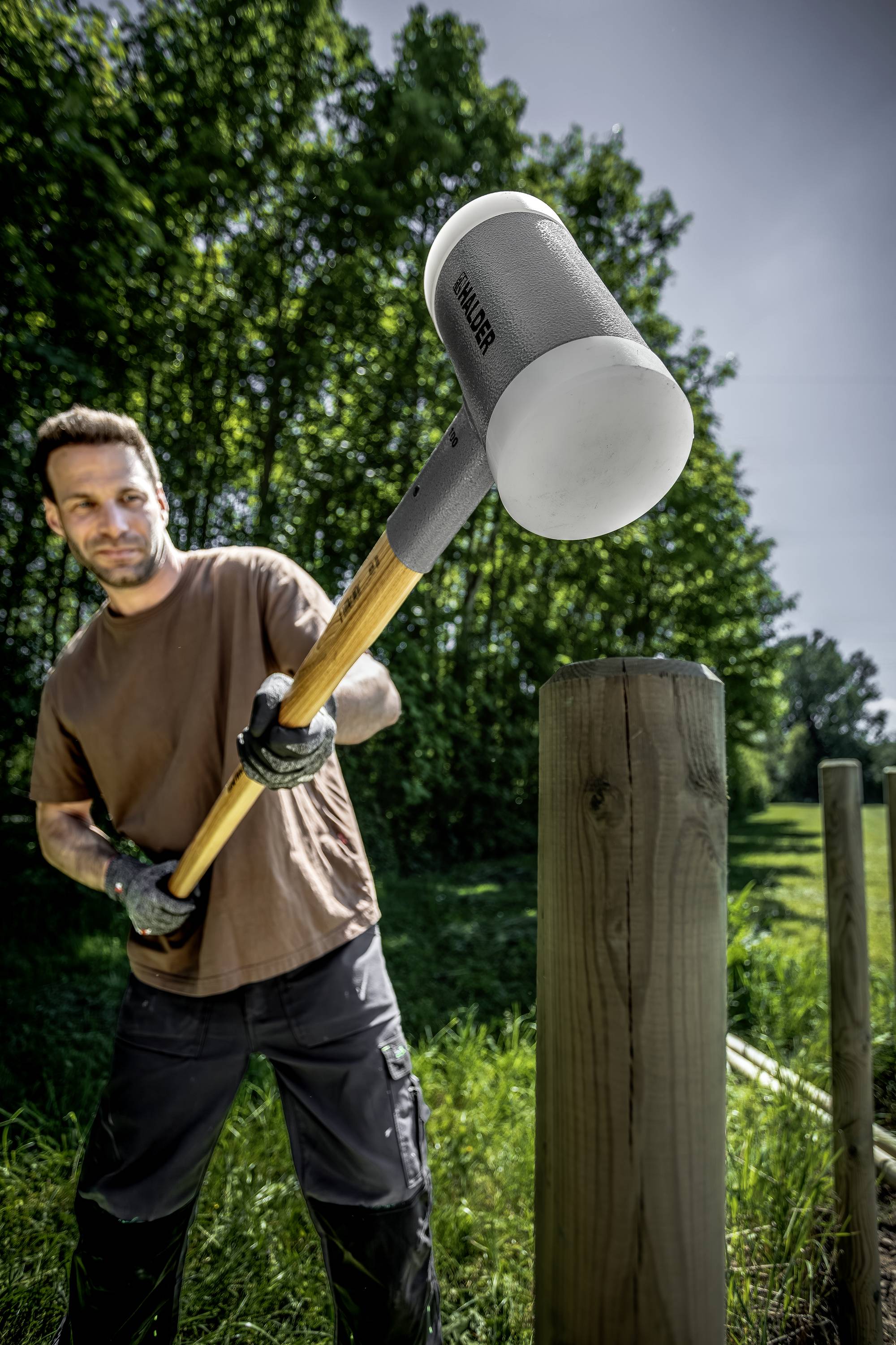 A man is hammering a large wooden post into the ground outdoors. Trees and grass can be seen in the background.