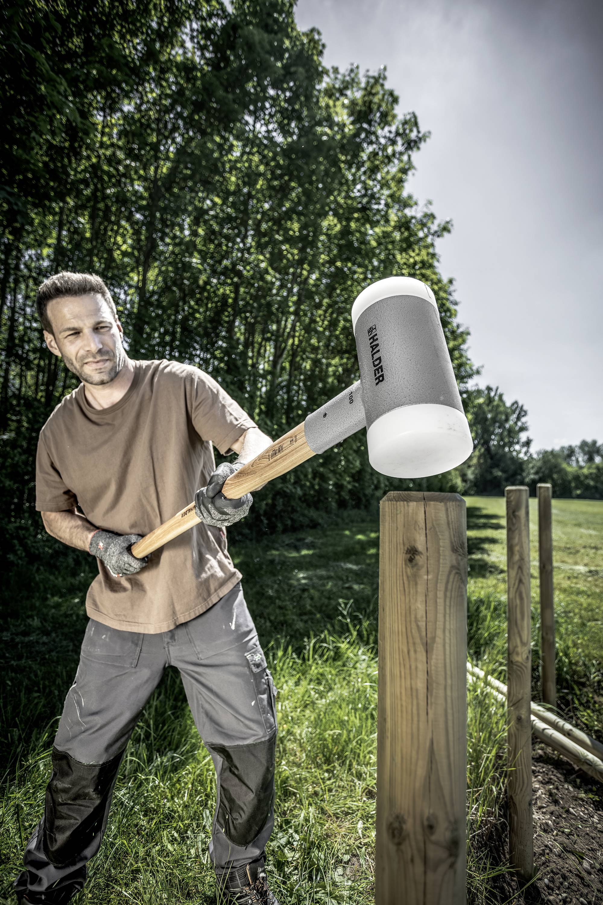 A man in workwear is hammering a wooden post into the ground with a large hammer to erect a fence in a green environment.