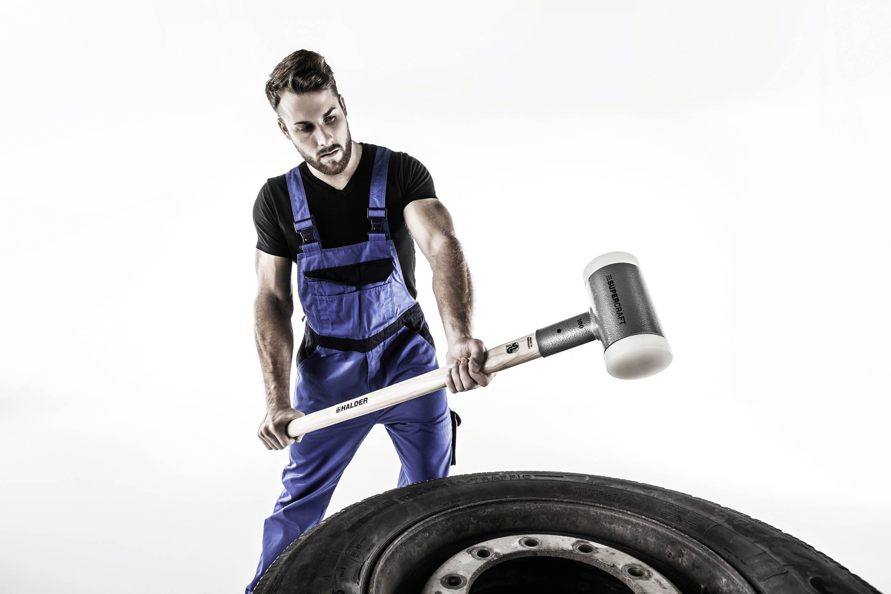 A man in blue workwear is using a large hammer to strike a car tyre.