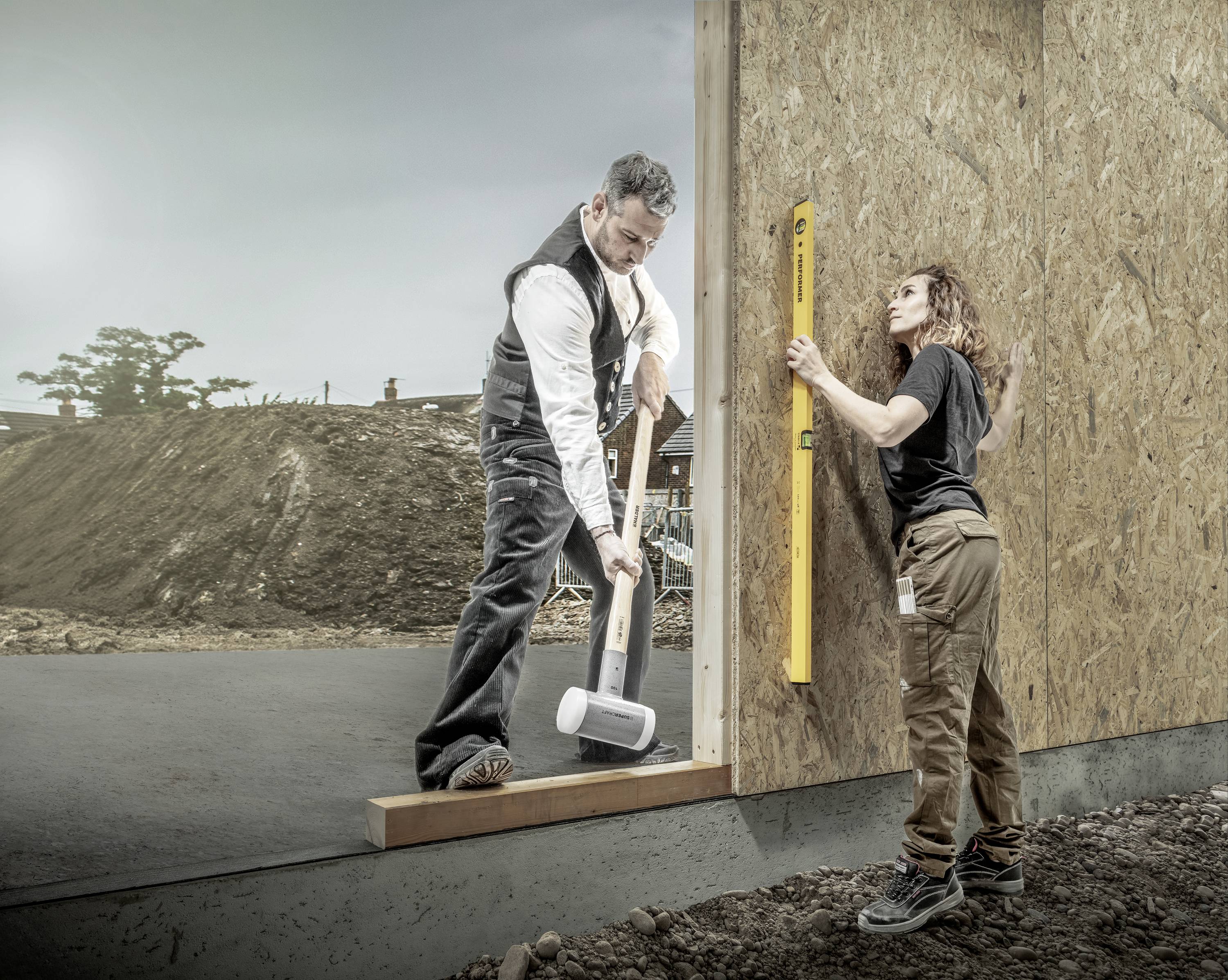 A man and a woman are working together on a wooden construction outdoors. The man is using a hammer, and the woman is holding a spirit level.