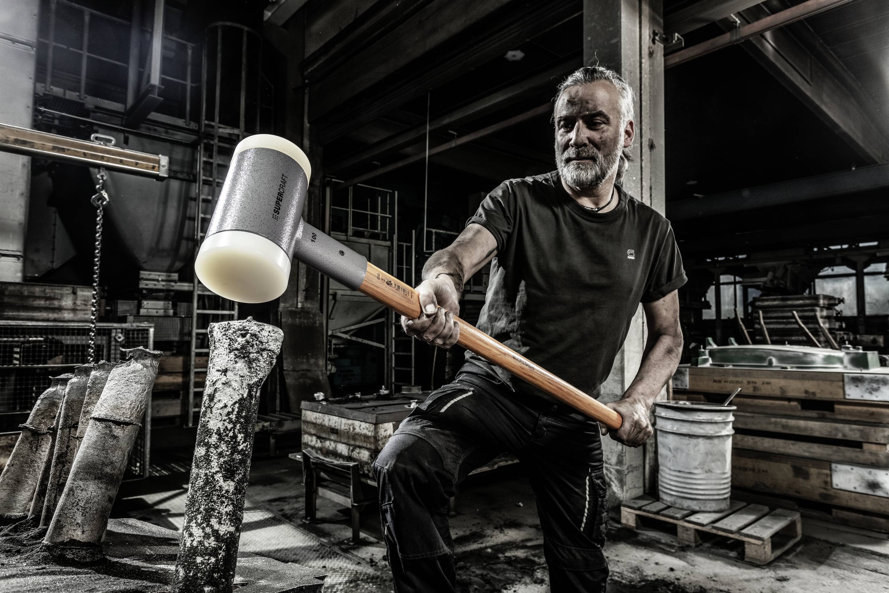A man is striking a stone block with a large hammer in an industrial workshop environment. He is wearing black workwear.