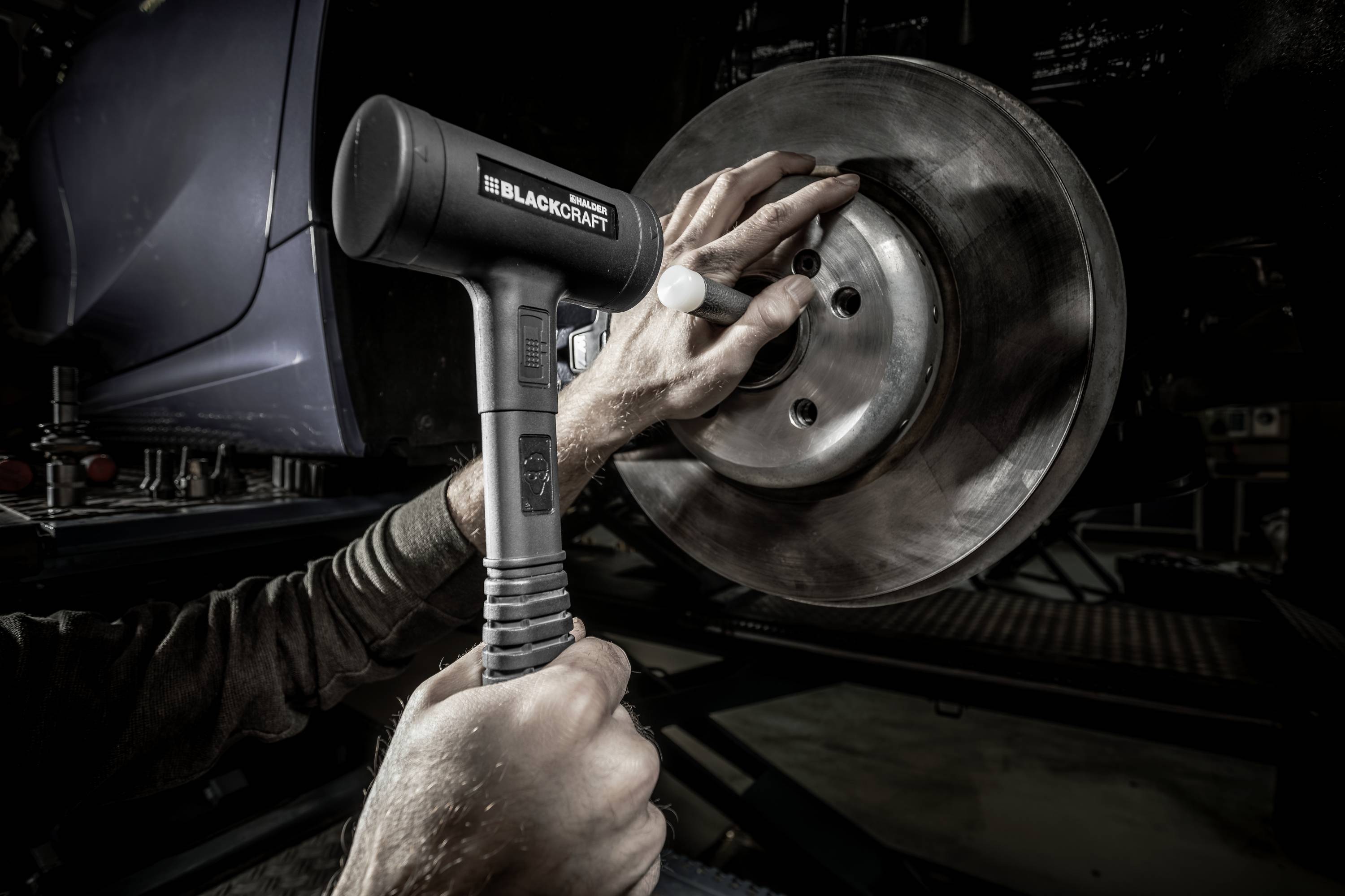 A person is using a tool to work on a brake disc of a car. Hands are close to the metal, the car is raised.