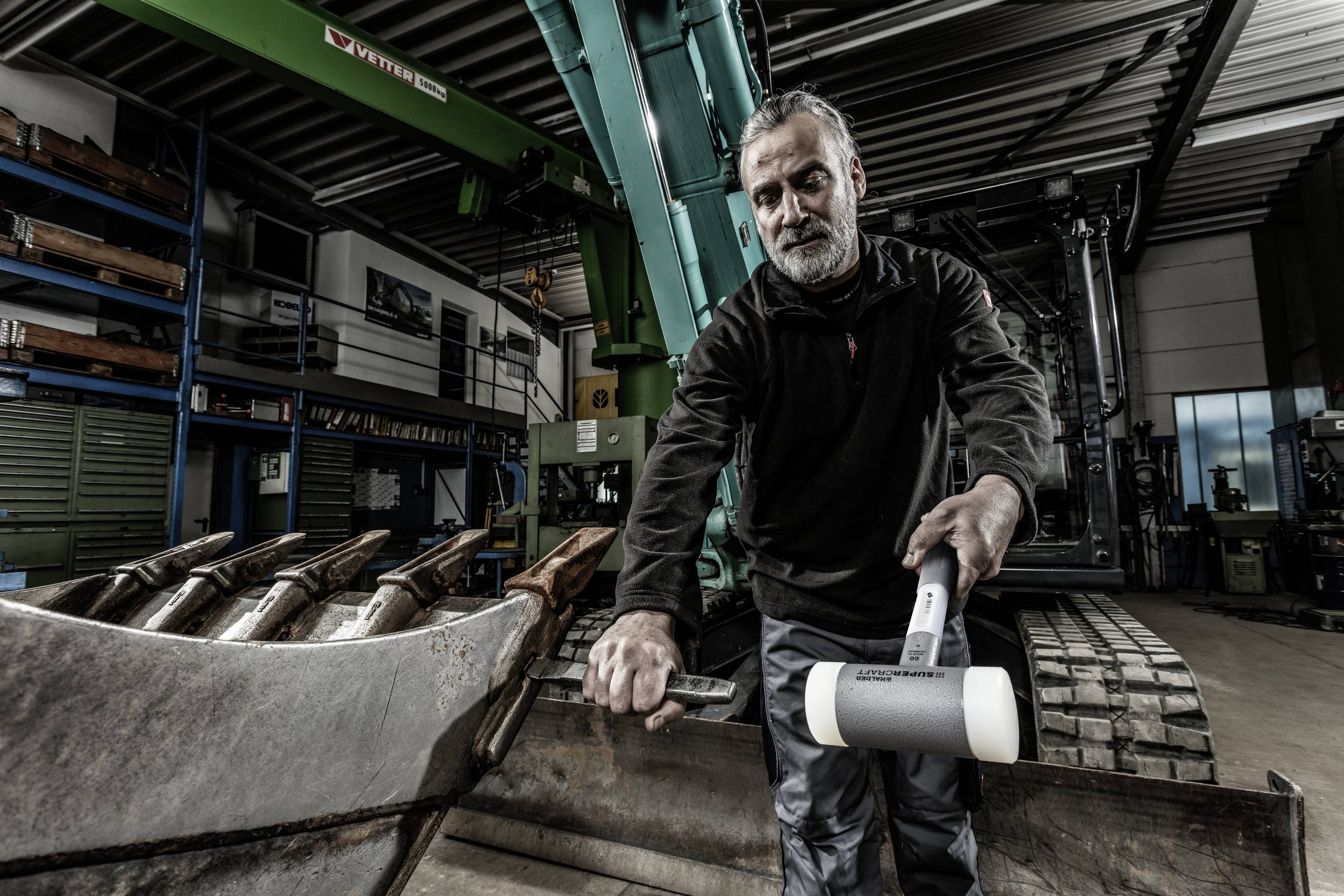 A man is standing in a workshop next to a digger, holding a white hammer. Shelves with tools can be seen in the background.