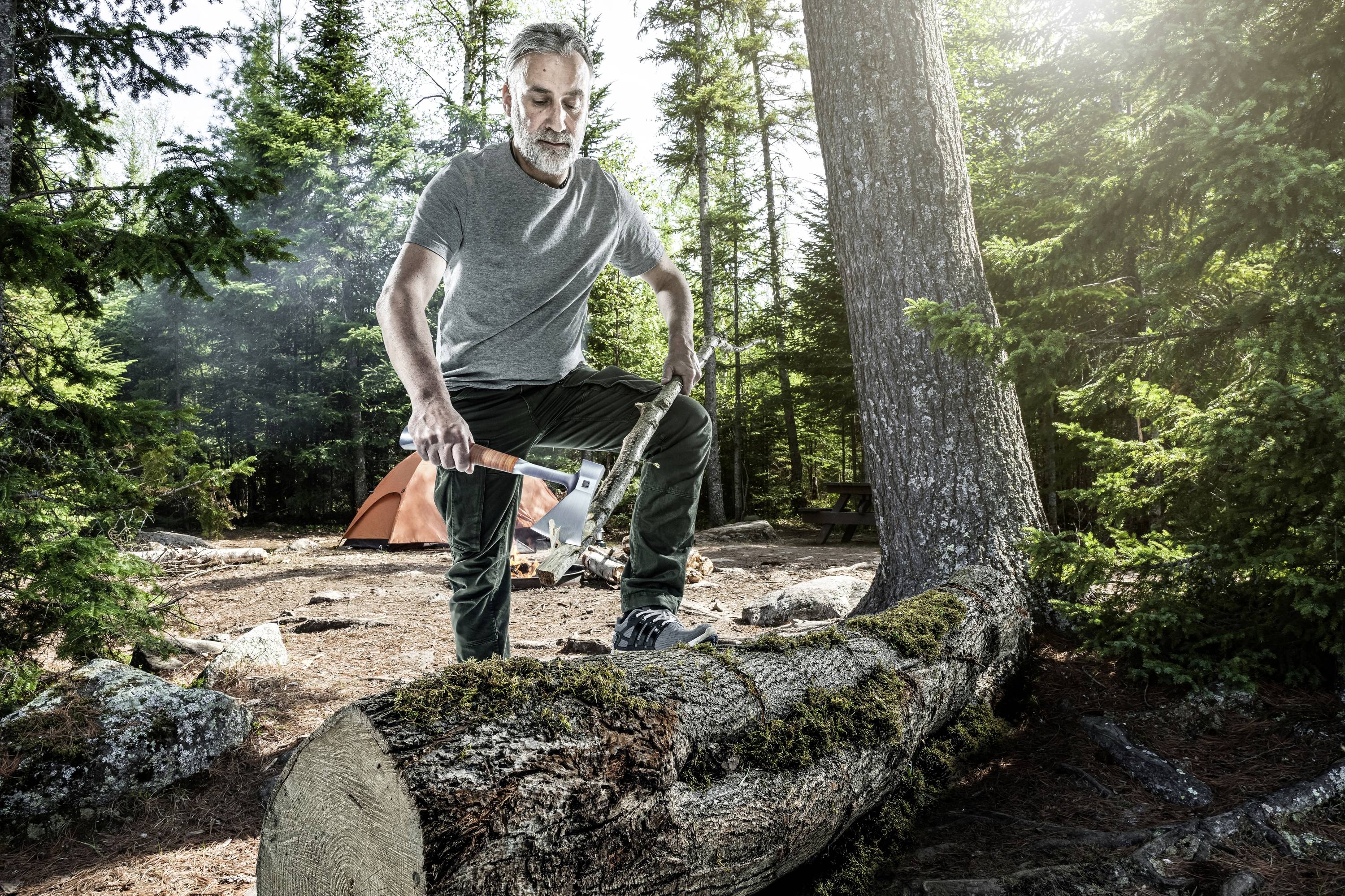 A man stands on a felled tree trunk, holding an axe. An orange tent is visible in the background.