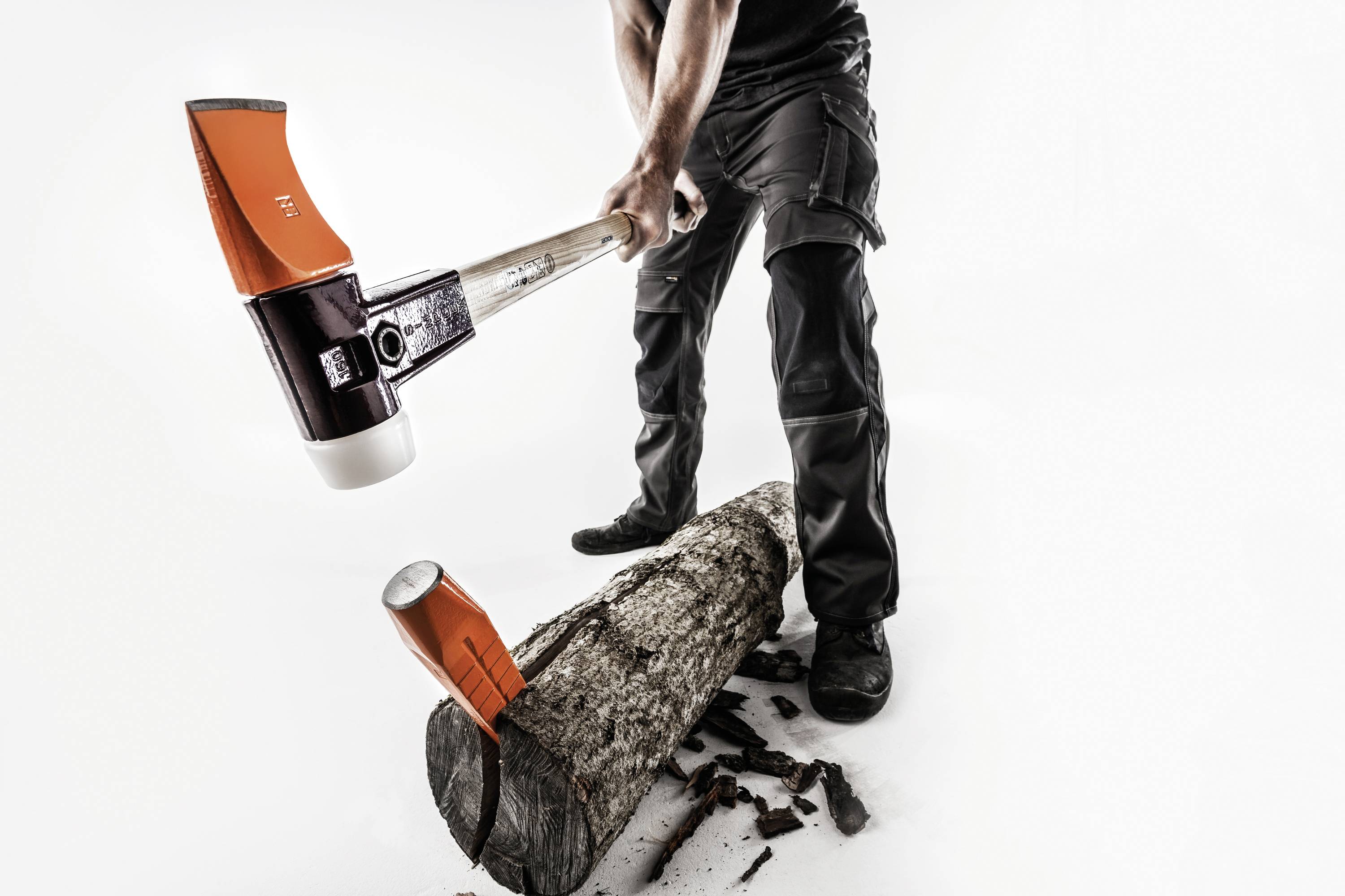 A person is splitting a tree trunk with an axe. Wood pieces are scattered on the ground. The background is white.