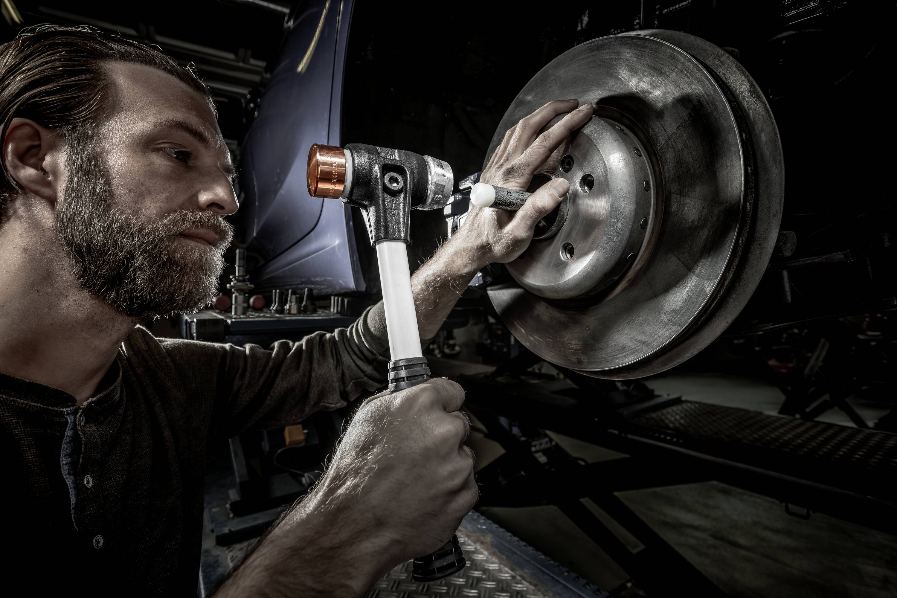 A man is using a pneumatic tool to work on a brake disc. Vehicle parts are visible in the background.