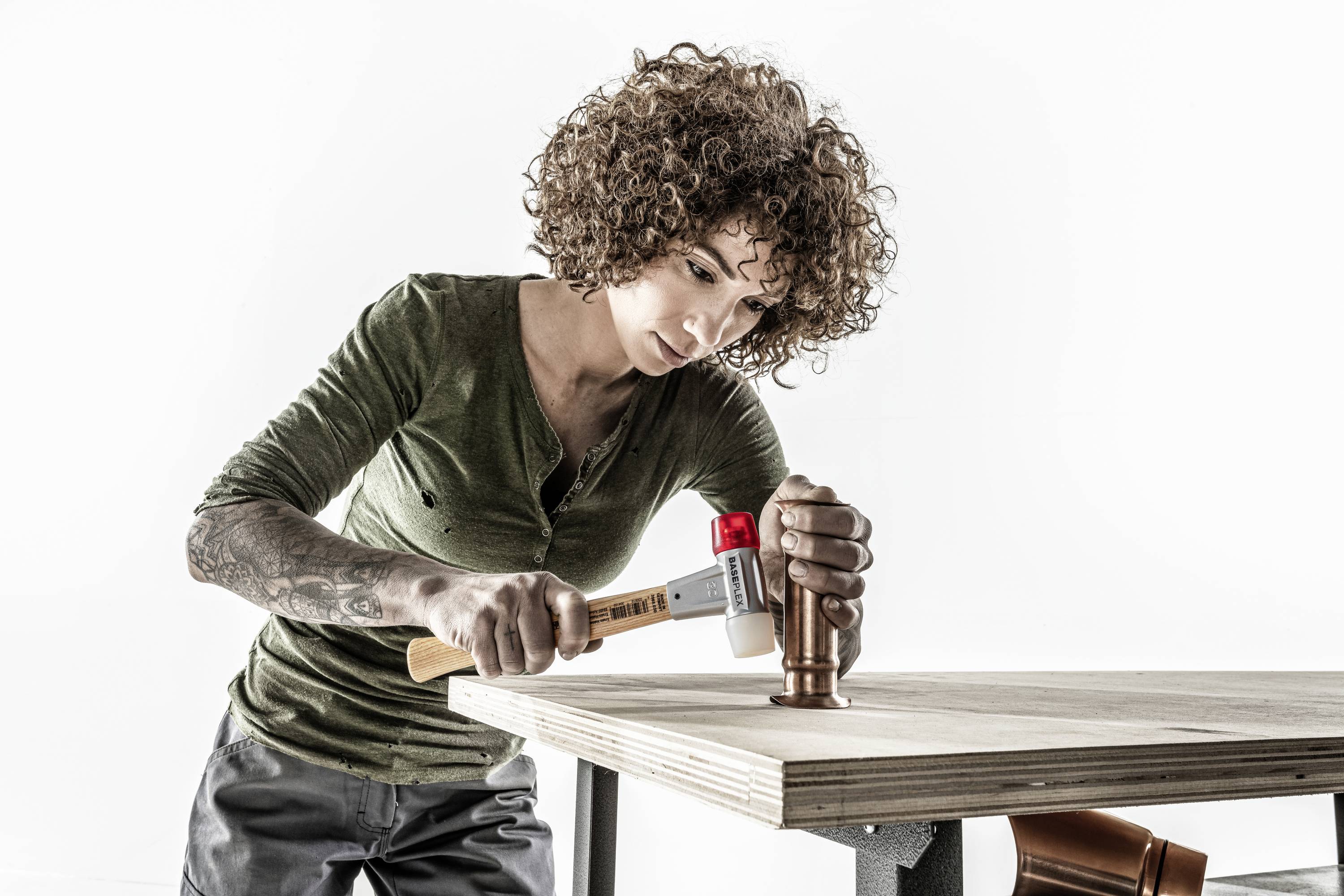 A person with curly hair is working intently with a hammer on a copper pipe on a wooden table.