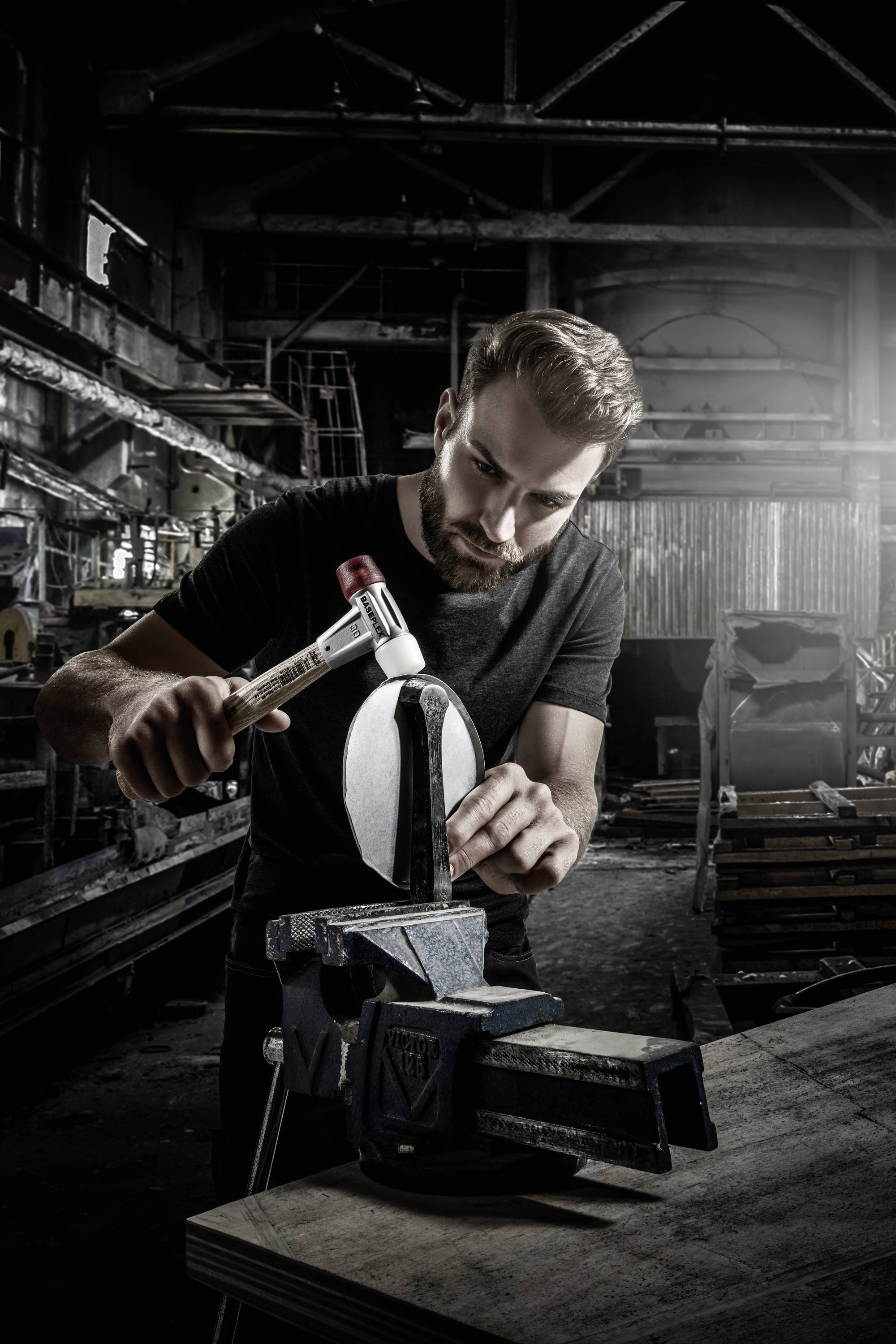 A man in a workshop is carefully hammering a piece of metal clamped in a vice. He is focused on his work.
