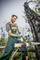 A man in workwear is felling a tree with an axe in a forest. The sky is clear, and already felled trees lie around him.