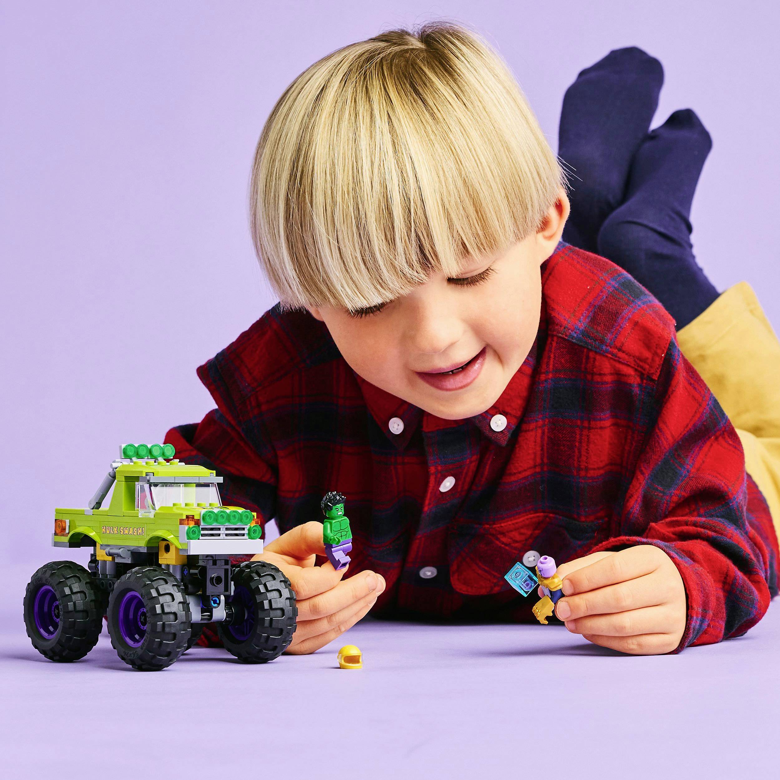 A child is playing on the floor with a toy car and figurines. They are wearing a checked shirt and lying on a yellow cushion.